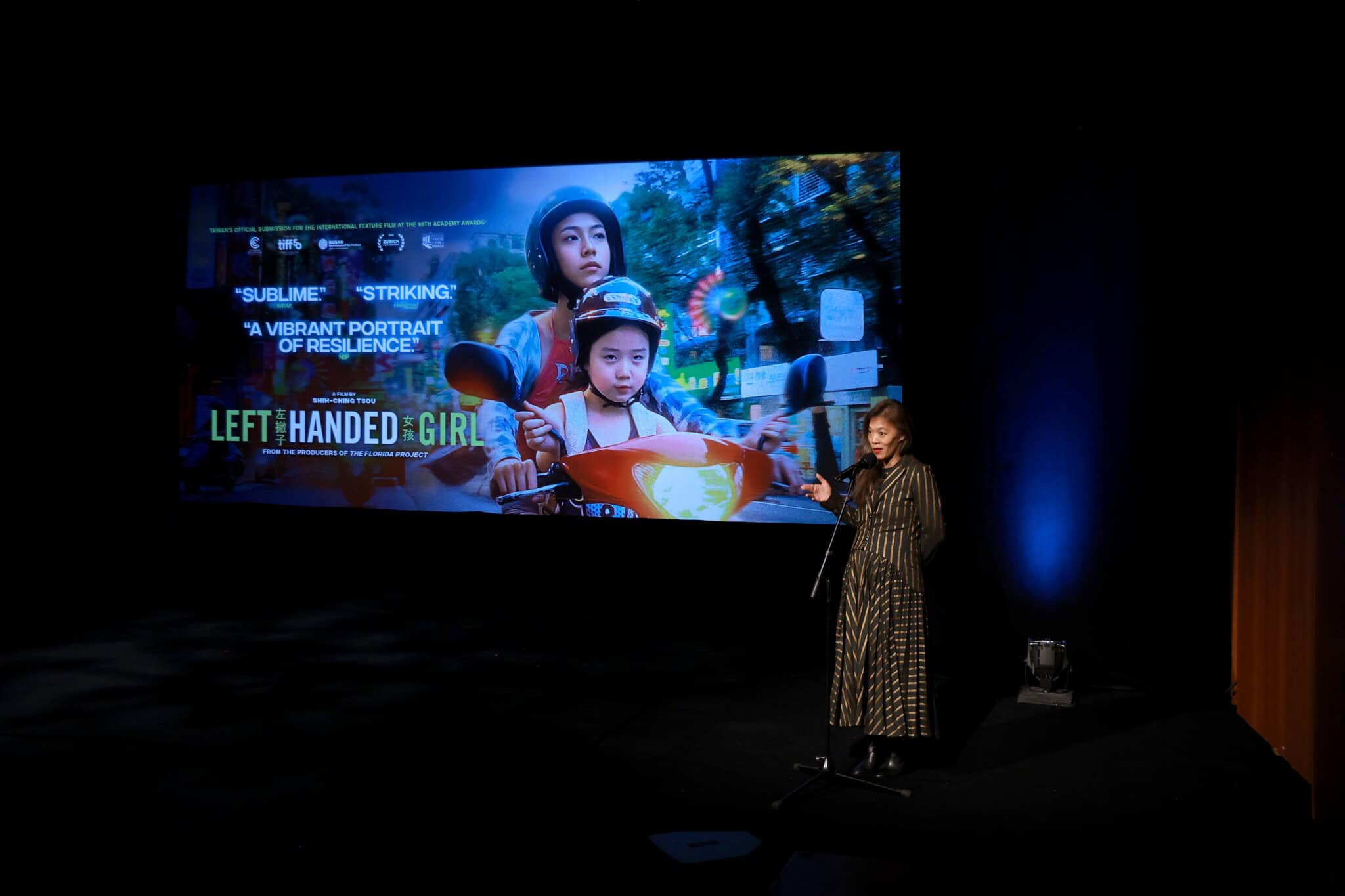 A woman stands on stage at a microphone, speaking in front of a large screen displaying a movie poster for Left-Handed Girl, featuring two girls riding a scooter. The stage is dimly lit with a spotlight on the speaker.
