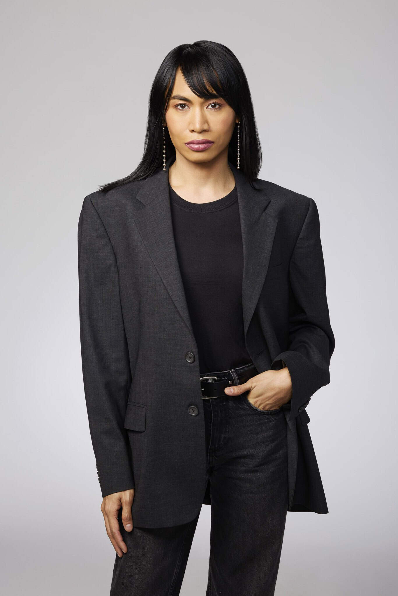 Person with long black hair wearing a black blazer, black shirt, and black jeans stands against a plain light gray background, looking directly at the camera with a neutral expression.