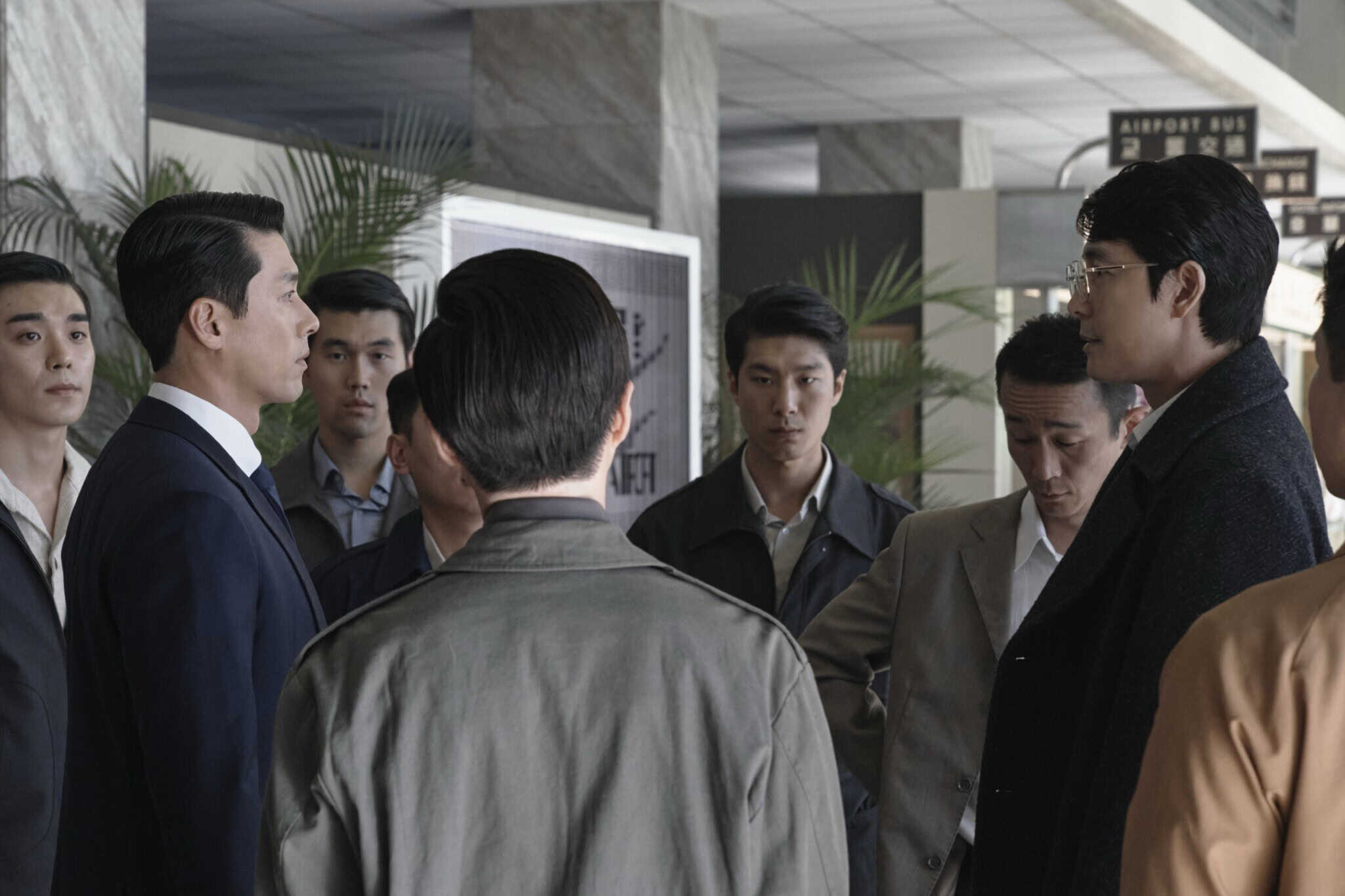 A group of men, some in suits and some in casual jackets, stand together indoors, facing each other in what appears to be a tense conversation. The setting looks like a public building or airport.