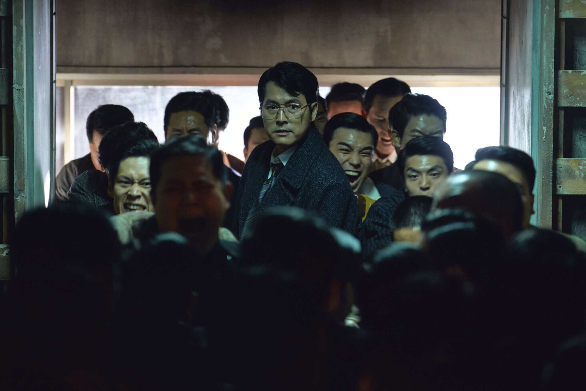 A serious man in a dark coat stands surrounded by a crowd of men in a dimly lit, crowded space, with intense expressions on several faces behind him.