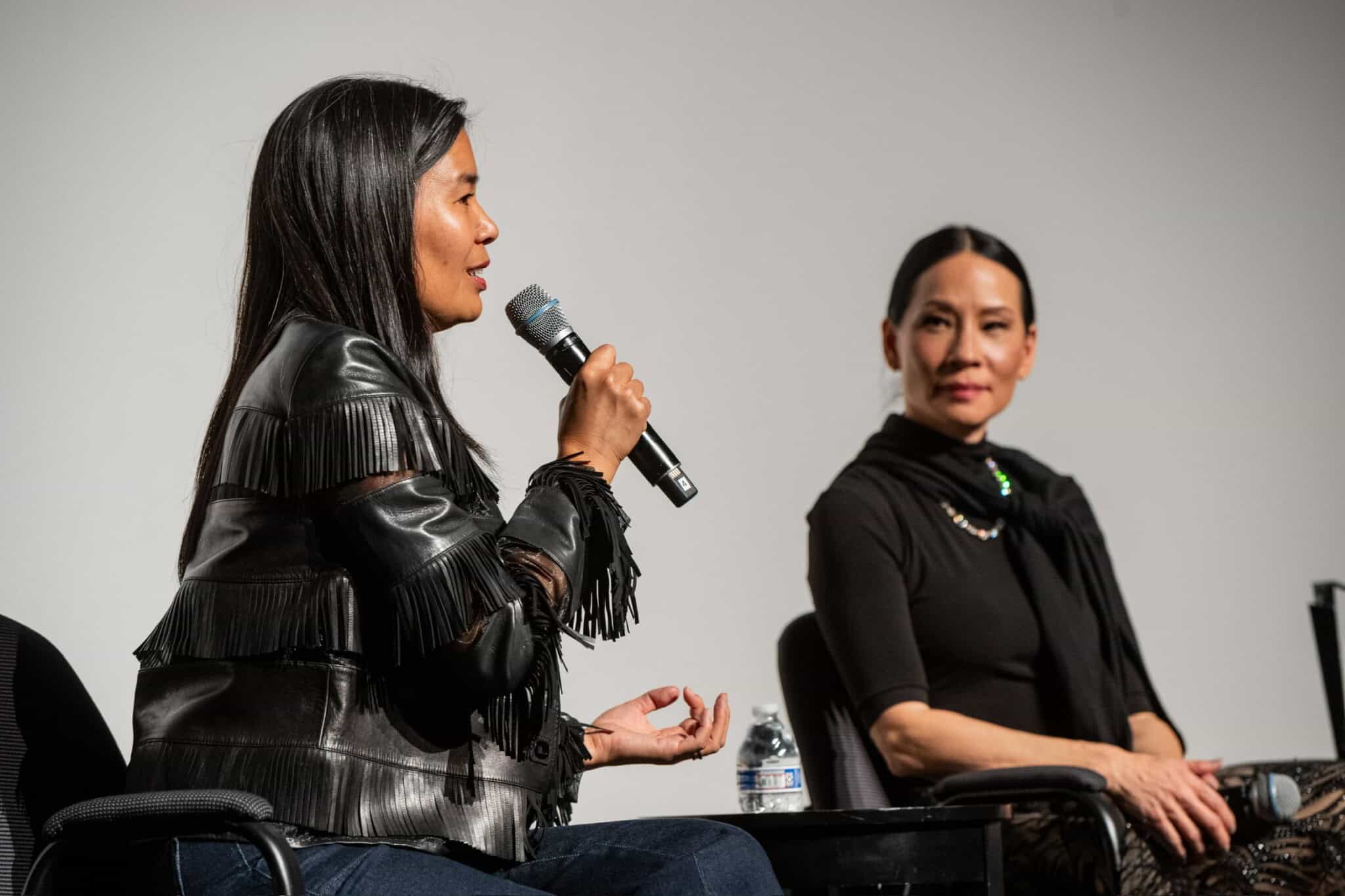 Two women sit on stage in conversation. One woman, holding a microphone and wearing a black fringed jacket, speaks while the other, dressed in black with a scarf, listens attentively.