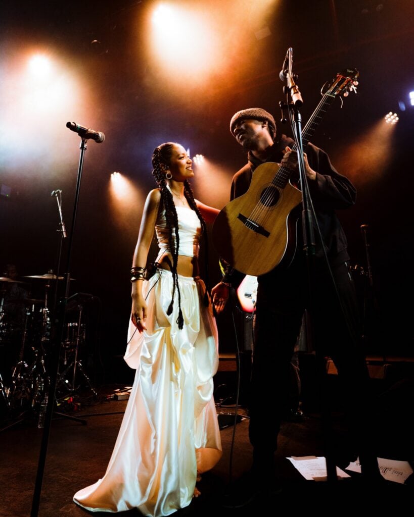 A woman in a white dress and a man holding an acoustic guitar stand close together on stage under warm, dramatic lighting, with microphones and musical equipment visible in the background.
