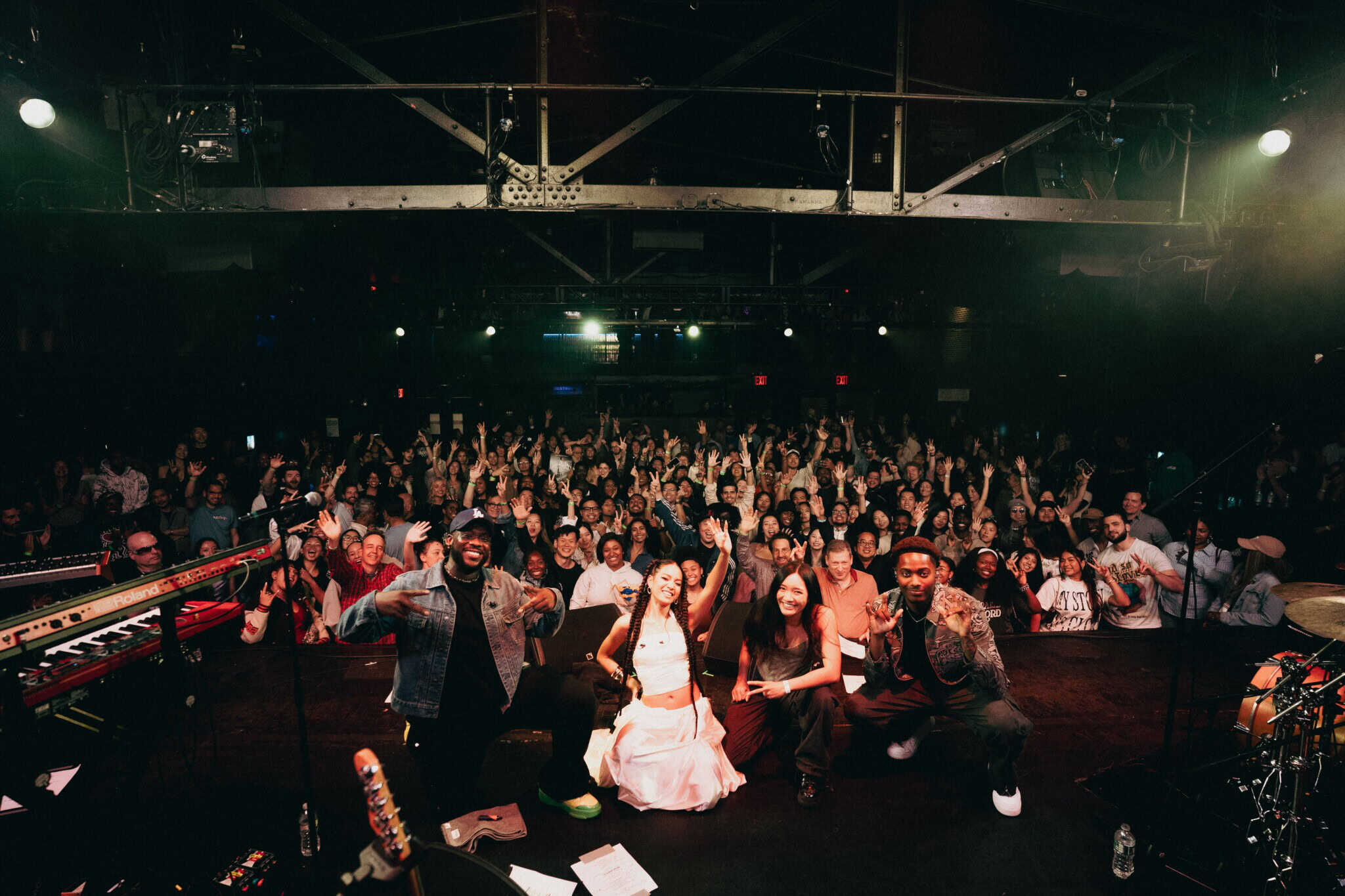 Four musicians pose and smile on stage with a large, cheering crowd behind them in a concert venue. Stage lights illuminate the band and the audience enthusiastically holds up their hands.