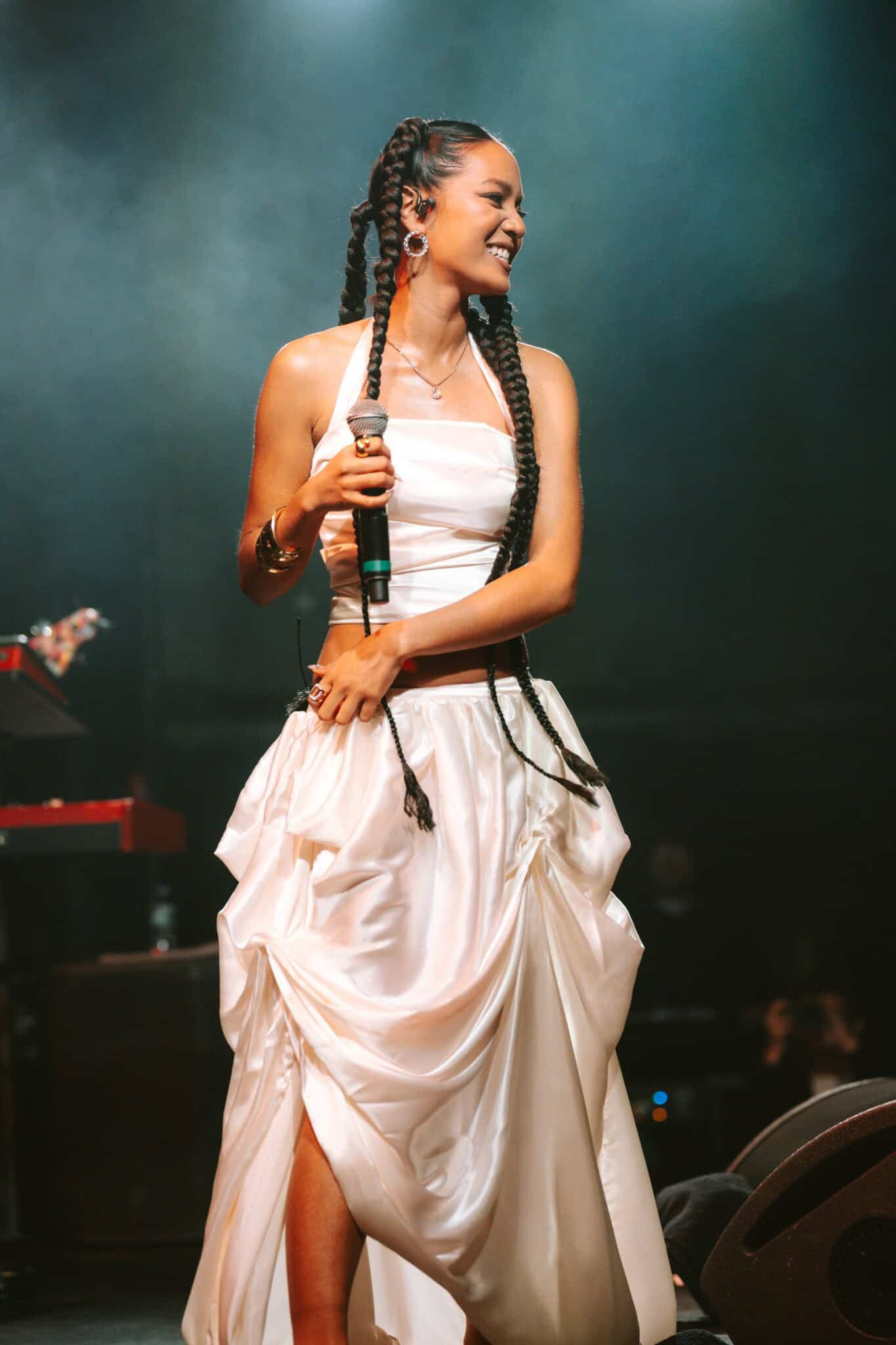 A woman with long braids and hoop earrings smiles while holding a microphone on stage. She wears a white halter top and a ruched white skirt. The background is dark with stage equipment visible.