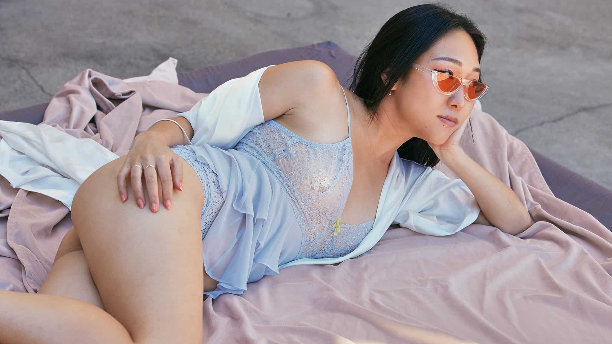A woman in pastel blue lingerie and a white robe lounges on fabric outdoors, wearing orange-tinted sunglasses and gazing into the distance with a relaxed expression.