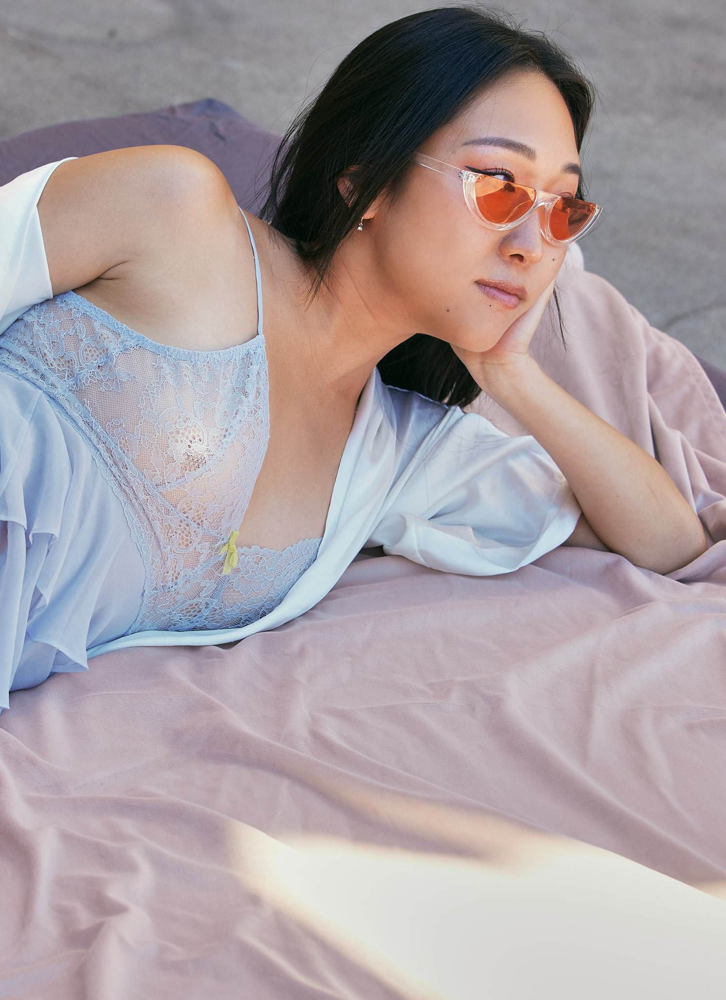 A woman in pastel blue lingerie and a white robe lounges on fabric outdoors, wearing orange-tinted sunglasses and gazing into the distance with a relaxed expression.