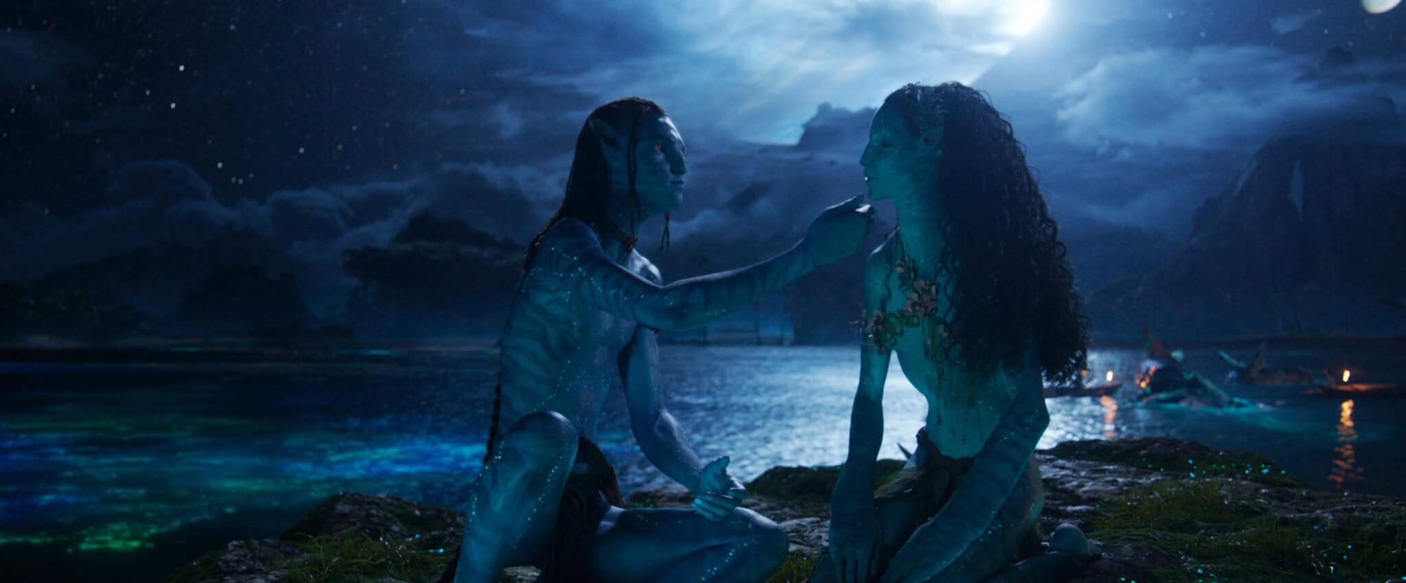 Two blue-skinned humanoid characters sit together by the water under a bright moon, with one gently touching the others chin. Lush scenery and glowing reflections create a mystical nighttime atmosphere.