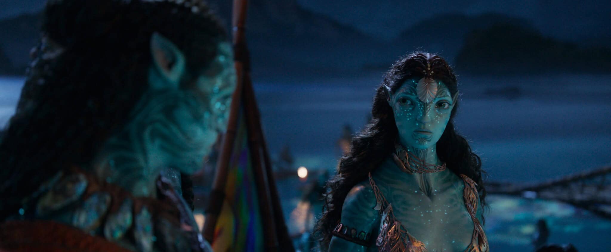 Two blue-skinned, humanoid alien characters with glowing spots stand facing each other at night near water, with mountains and soft lights in the background. One character looks intently at the other.