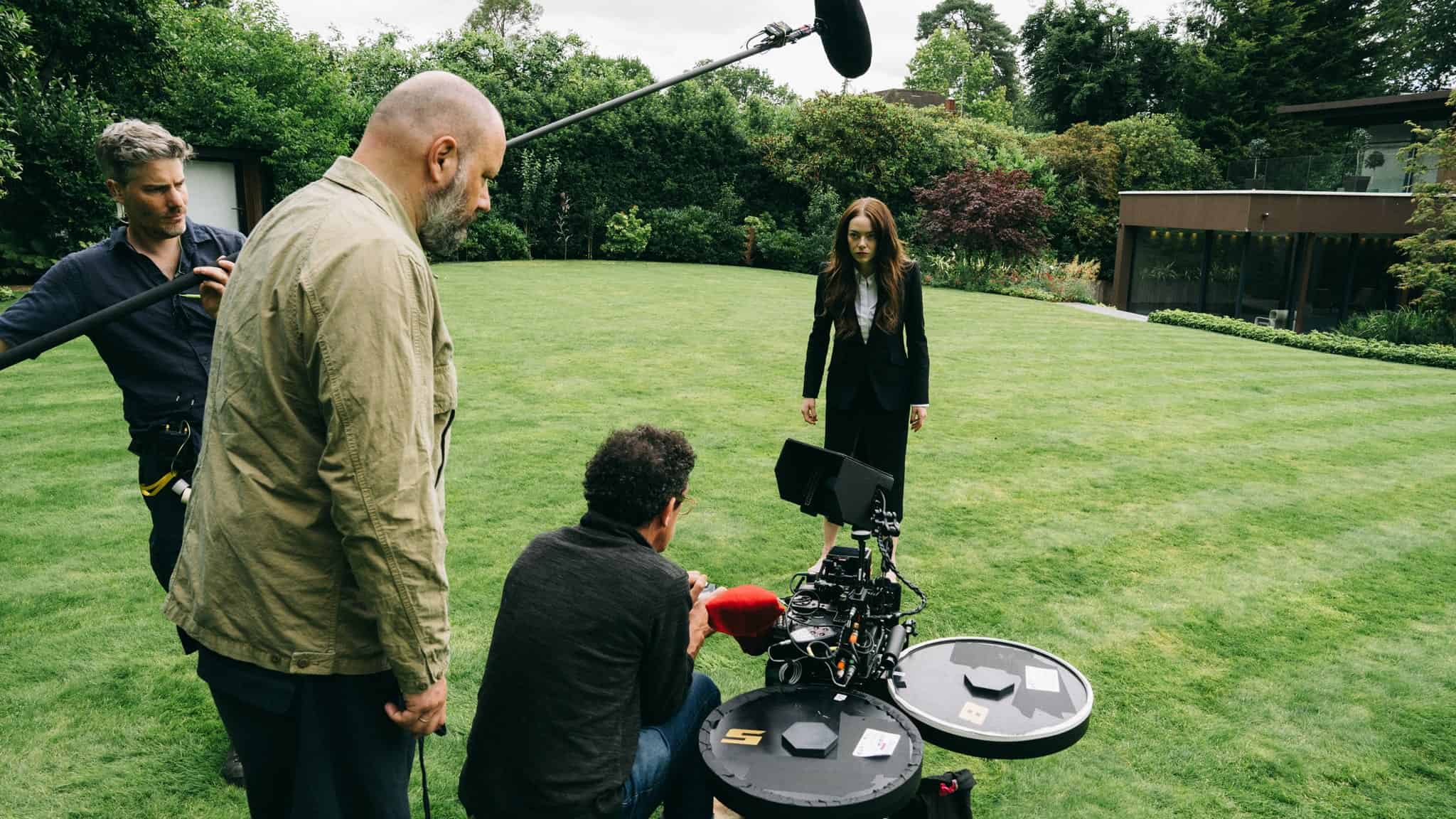 A film crew sets up camera equipment and a boom mic on a grassy lawn while a woman in a black suit stands in front of them, preparing for a scene. Trees and a modern building are in the background.
