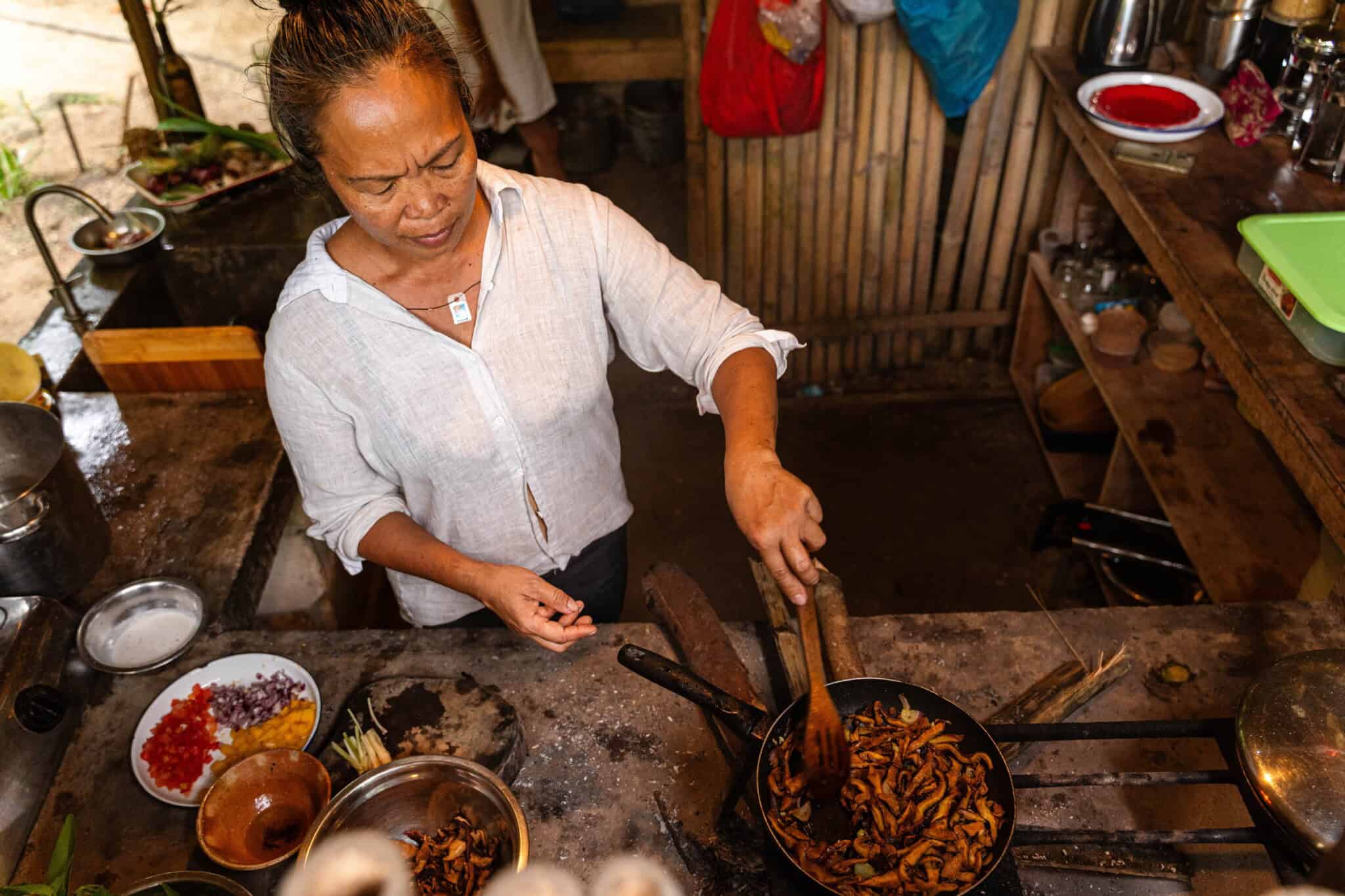 A woman in a white shirt cooks mushrooms in a pan inside a rustic kitchen with wooden shelves, utensils, and ingredients around her. She is using tongs to stir the food on the stove.