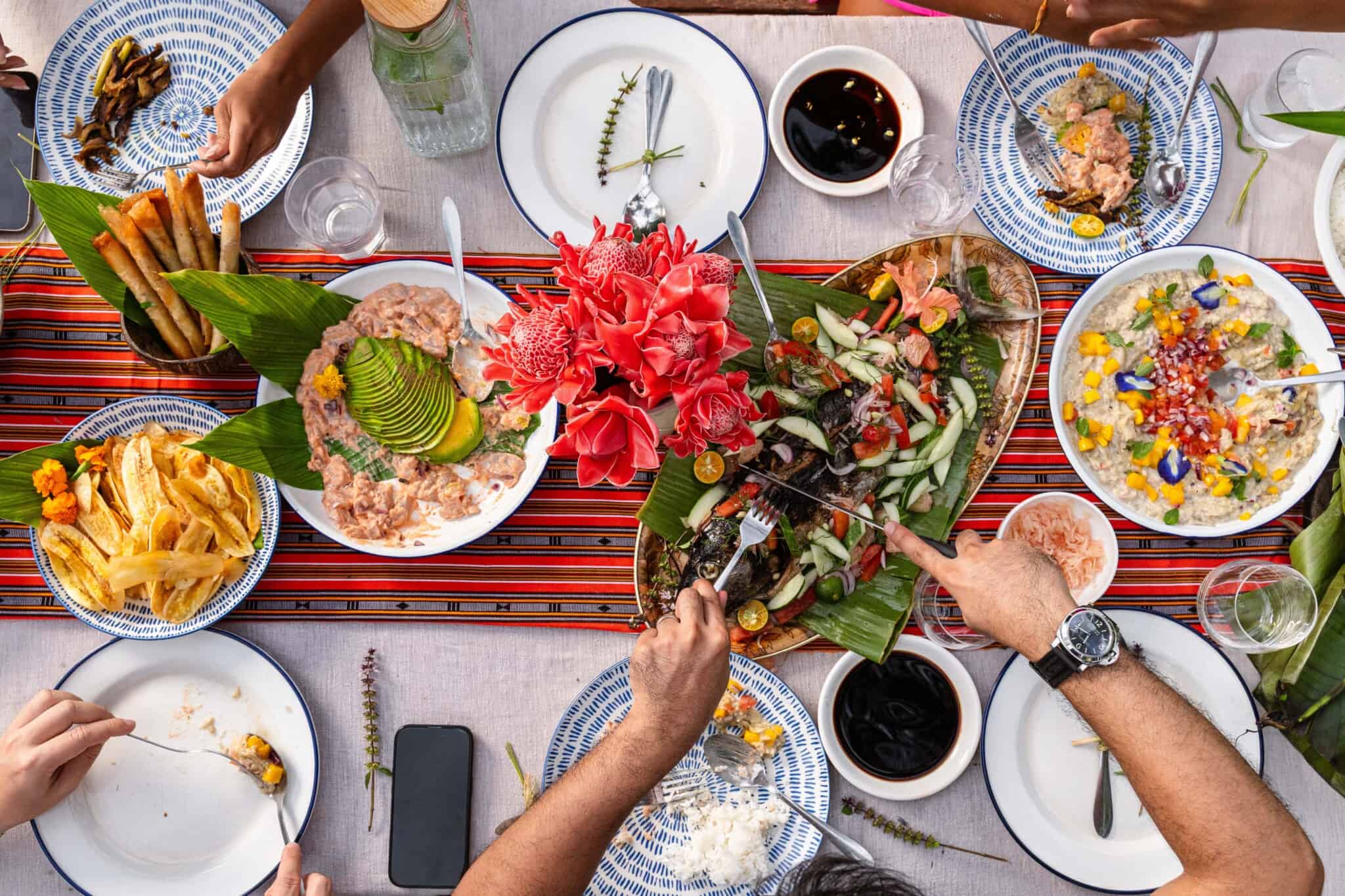 A colorful top-down view of a table set with various vibrant Filipino dishes, plates, drinks, and a centerpiece of red flowers, as several people reach in to serve themselves and share food.