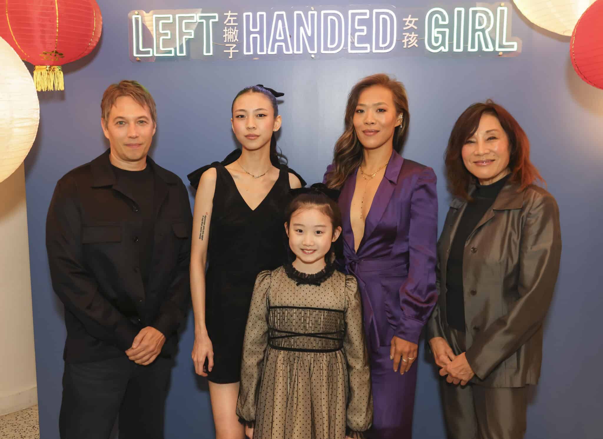 Five people stand together in front of a neon sign reading Left Handed Girl, with red and white lanterns on each side. Four adults and one child are dressed in semi-formal attire, smiling at the camera.