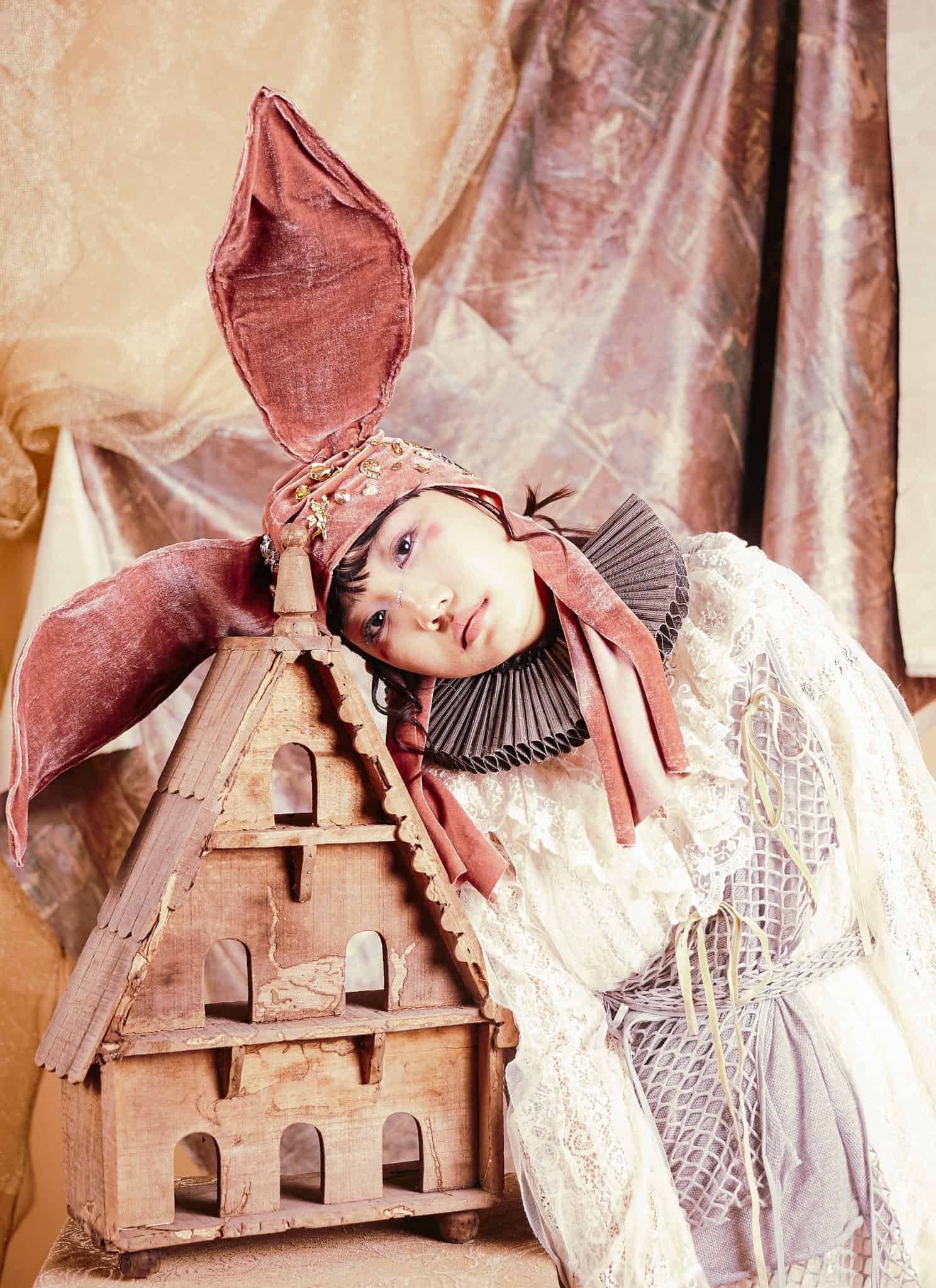 A person in whimsical, vintage-style clothing with a large pink bunny ear headpiece leans their head on a wooden birdhouse against a soft, draped fabric background.