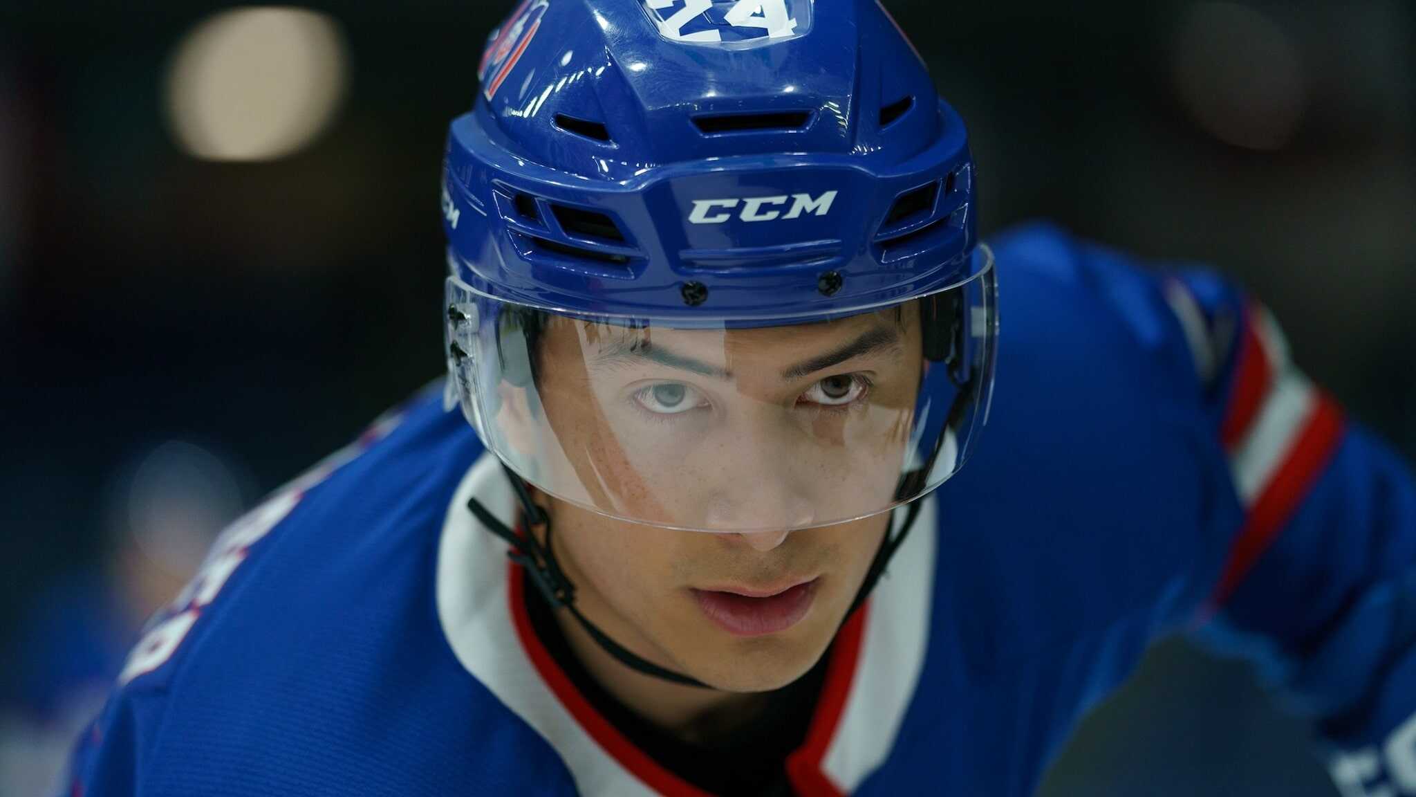 A close-up of a focused ice hockey player wearing a blue helmet and jersey, with a clear visor covering his face, looking intently ahead on the ice.