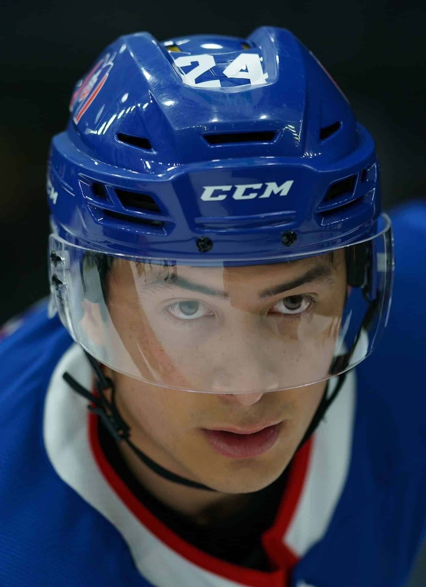 A close-up of a focused ice hockey player wearing a blue helmet and jersey, with a clear visor covering his face, looking intently ahead on the ice.