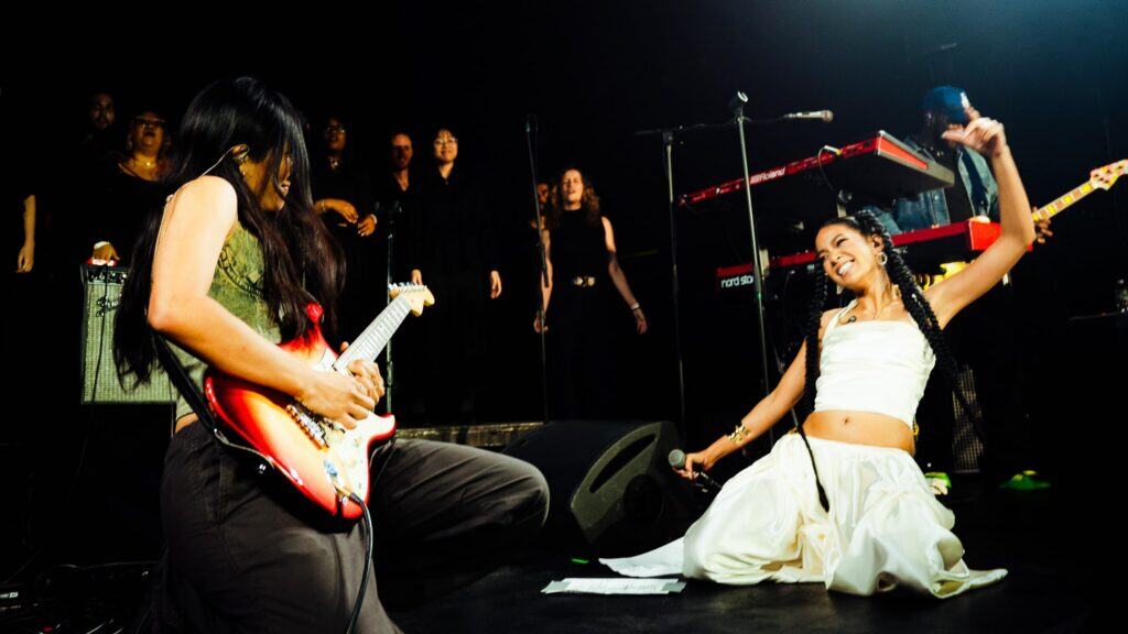 Two musicians perform energetically on stage; one plays electric guitar while kneeling, the other, dressed in white, sits on the floor singing passionately. A backup group and musical equipment are visible in the background.