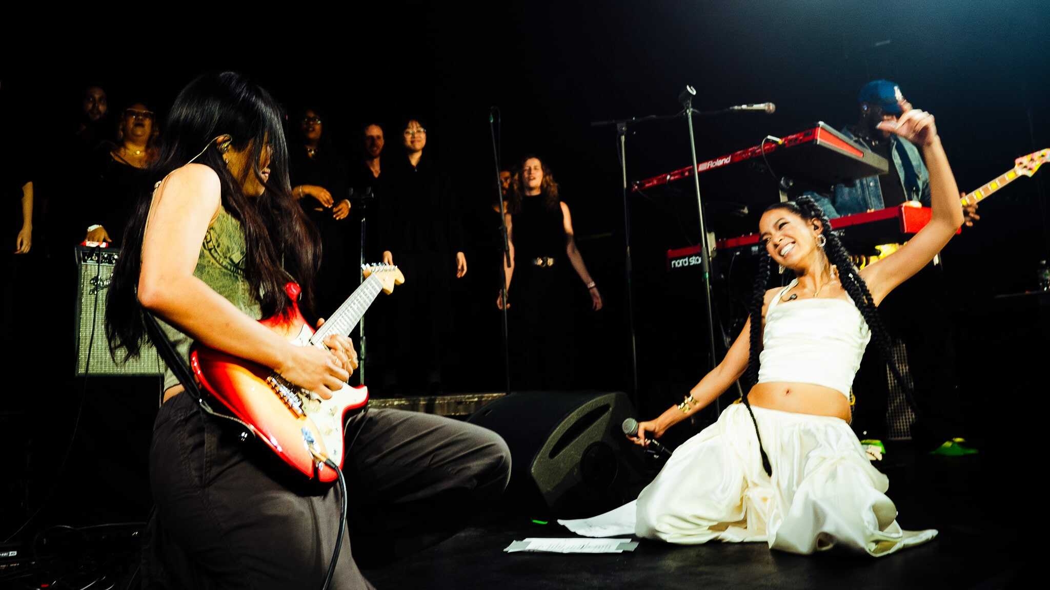 Two musicians perform energetically on stage; one plays electric guitar while kneeling, the other, dressed in white, sits on the floor singing passionately. A backup group and musical equipment are visible in the background.