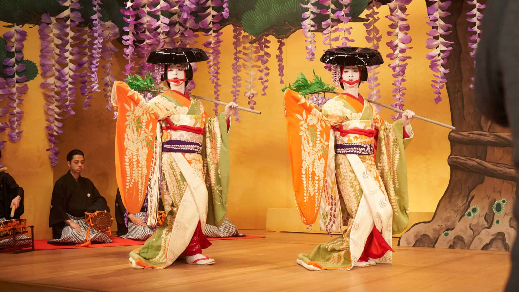 Two women in elaborate traditional Japanese kimonos and holding black umbrellas perform a dance on stage, surrounded by hanging purple wisteria flowers, with musicians in black robes seated in the background.