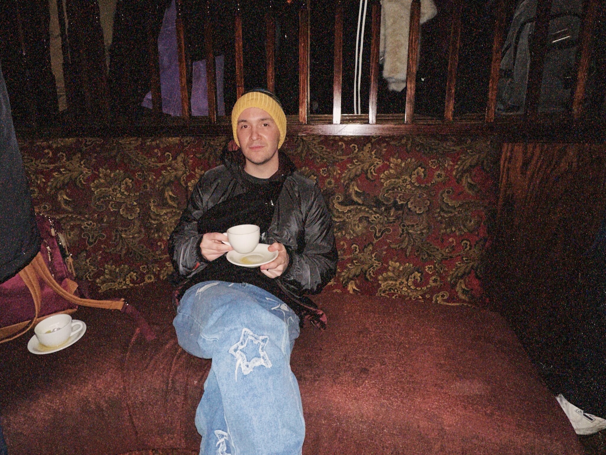 A person wearing a yellow beanie, black jacket, and ripped jeans sits on a patterned couch, holding a teacup and saucer, with legs crossed and a relaxed expression. A maroon bag and another teacup are nearby.