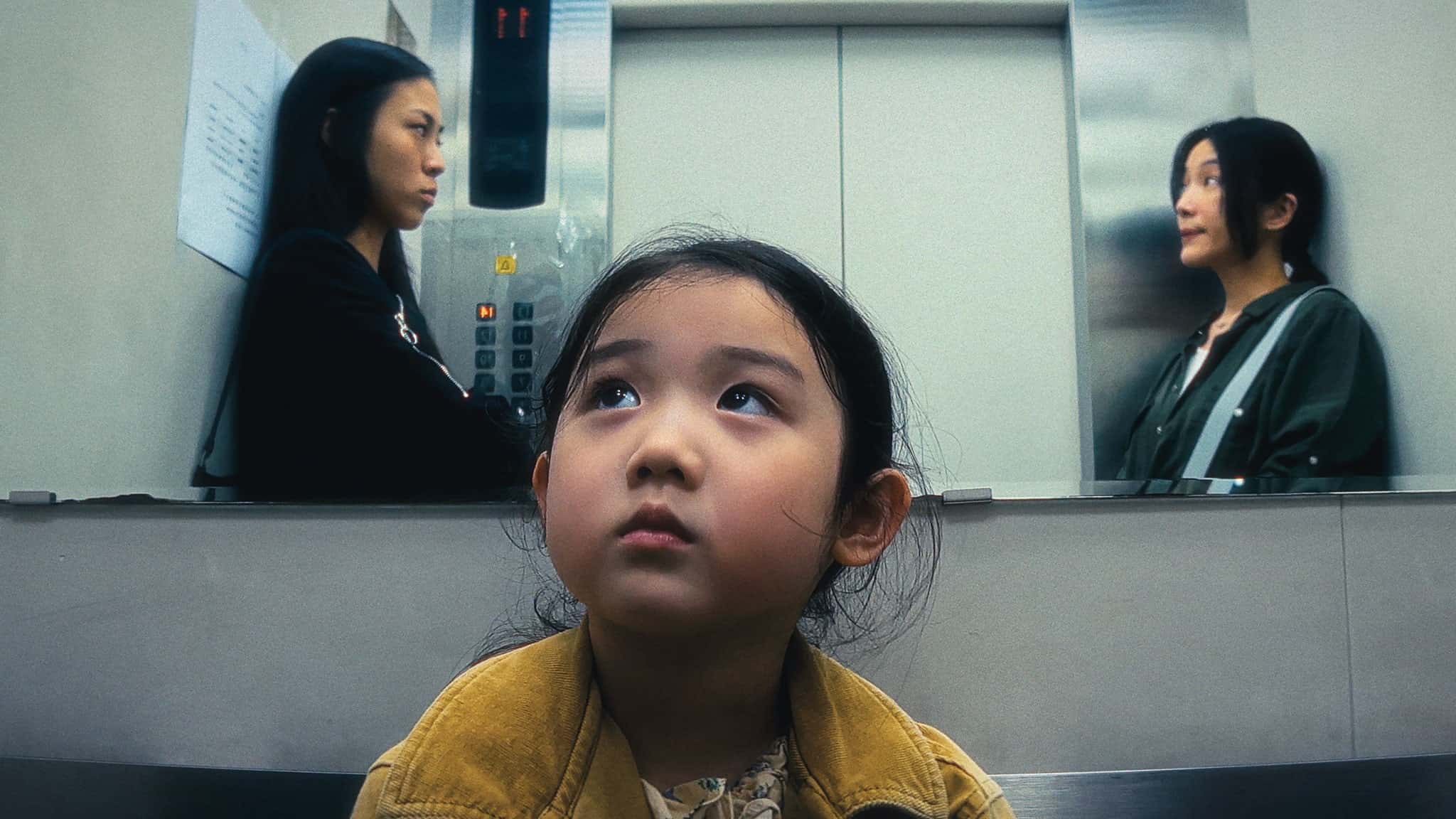 A young girl sits in the foreground of an elevator, looking up thoughtfully. Two women stand behind her on either side, facing each other with reflective expressions, each positioned next to a mirrored wall.