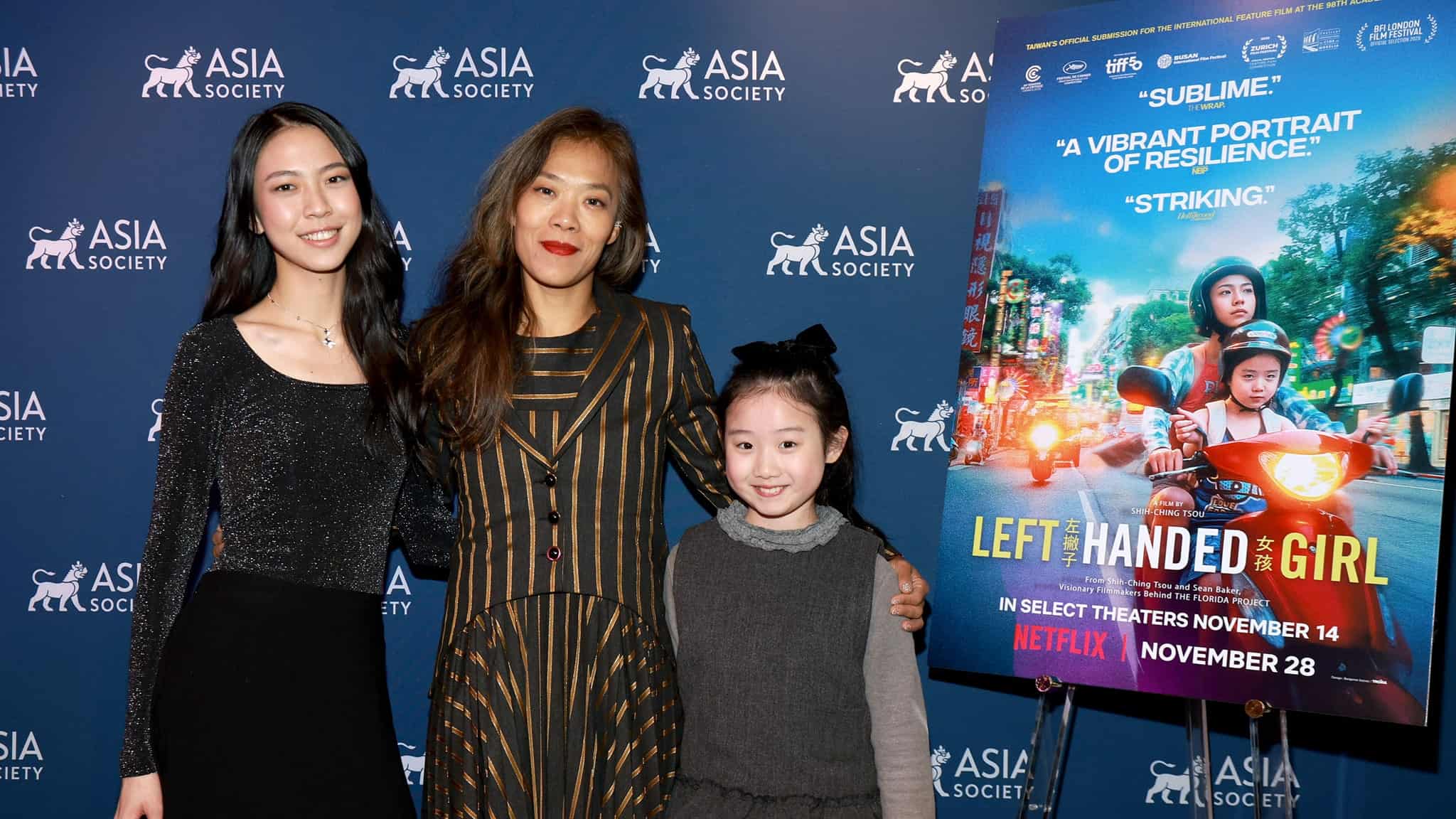 Three women stand smiling in front of an Asia Society backdrop next to a poster for the film Left-Handed Girl, which features a colorful scooter scene. The event promotes the films theatrical and Netflix release dates.
