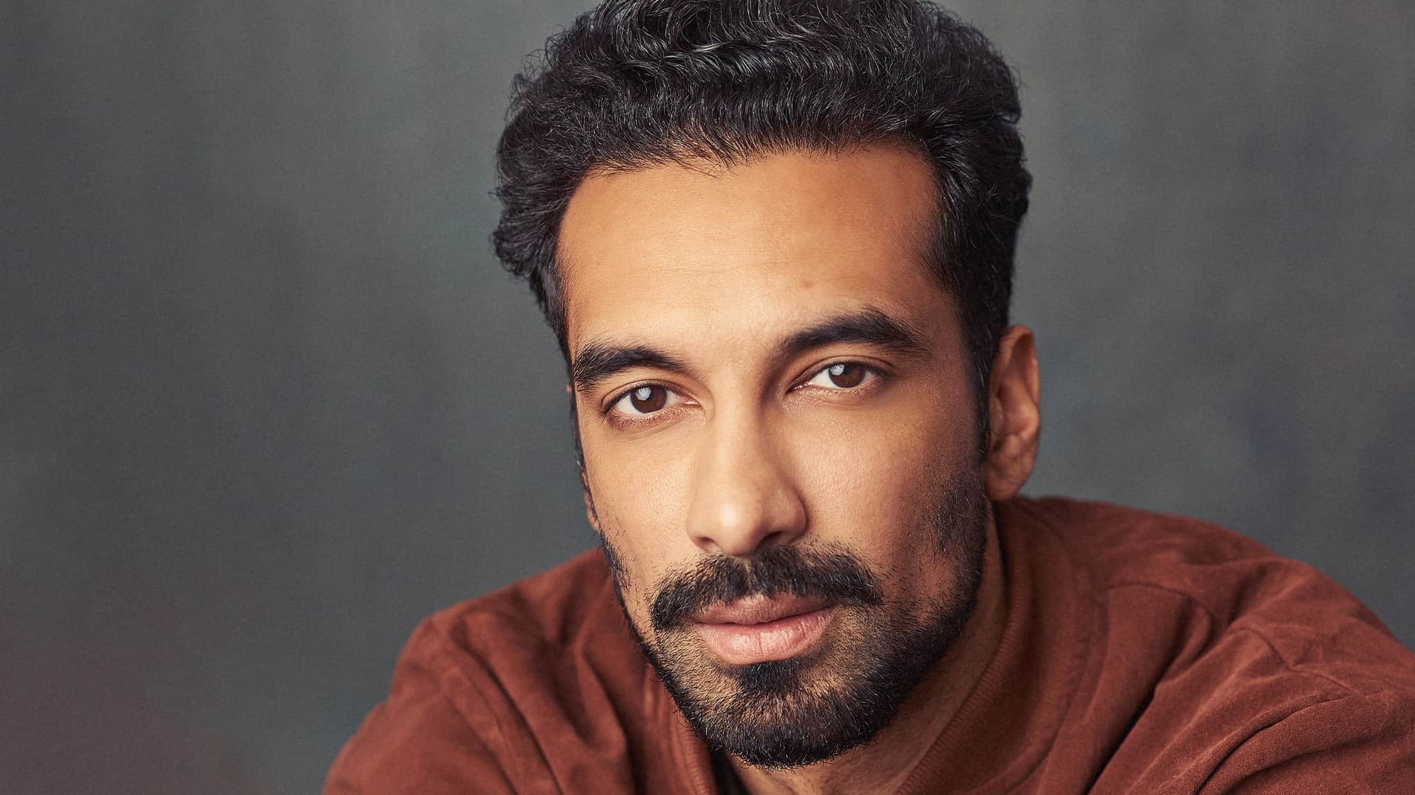 A man with short, dark curly hair and a trimmed beard wears a brown jacket and gazes directly at the camera, set against a blurred gray background.