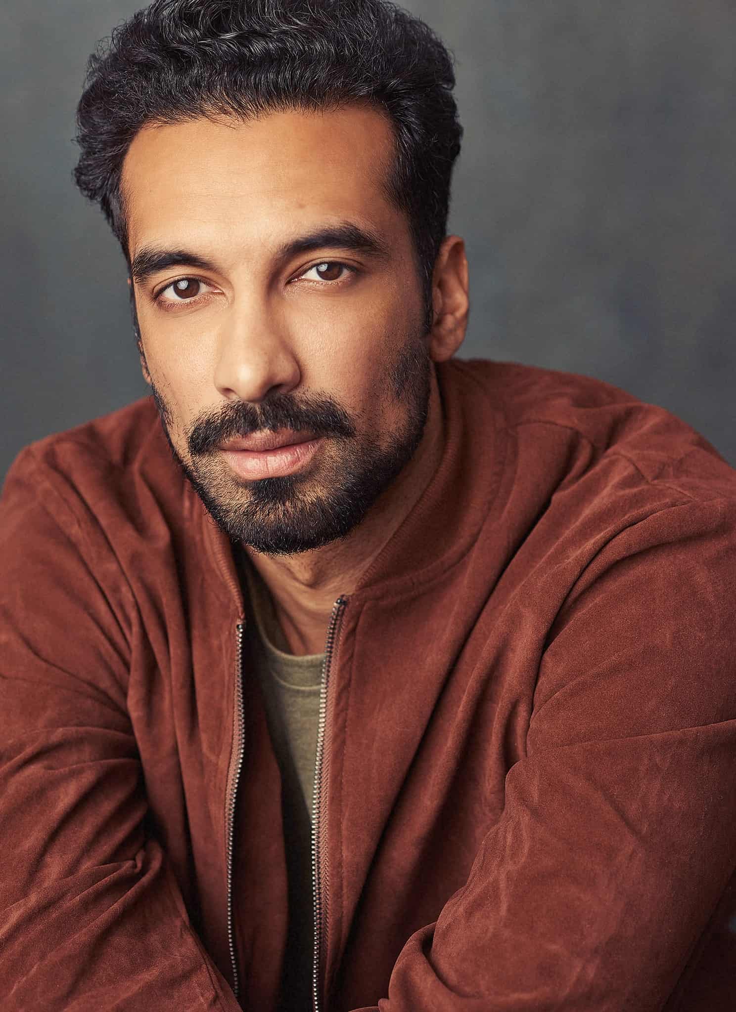 A man with short, dark curly hair and a trimmed beard wears a brown jacket and gazes directly at the camera, set against a blurred gray background.