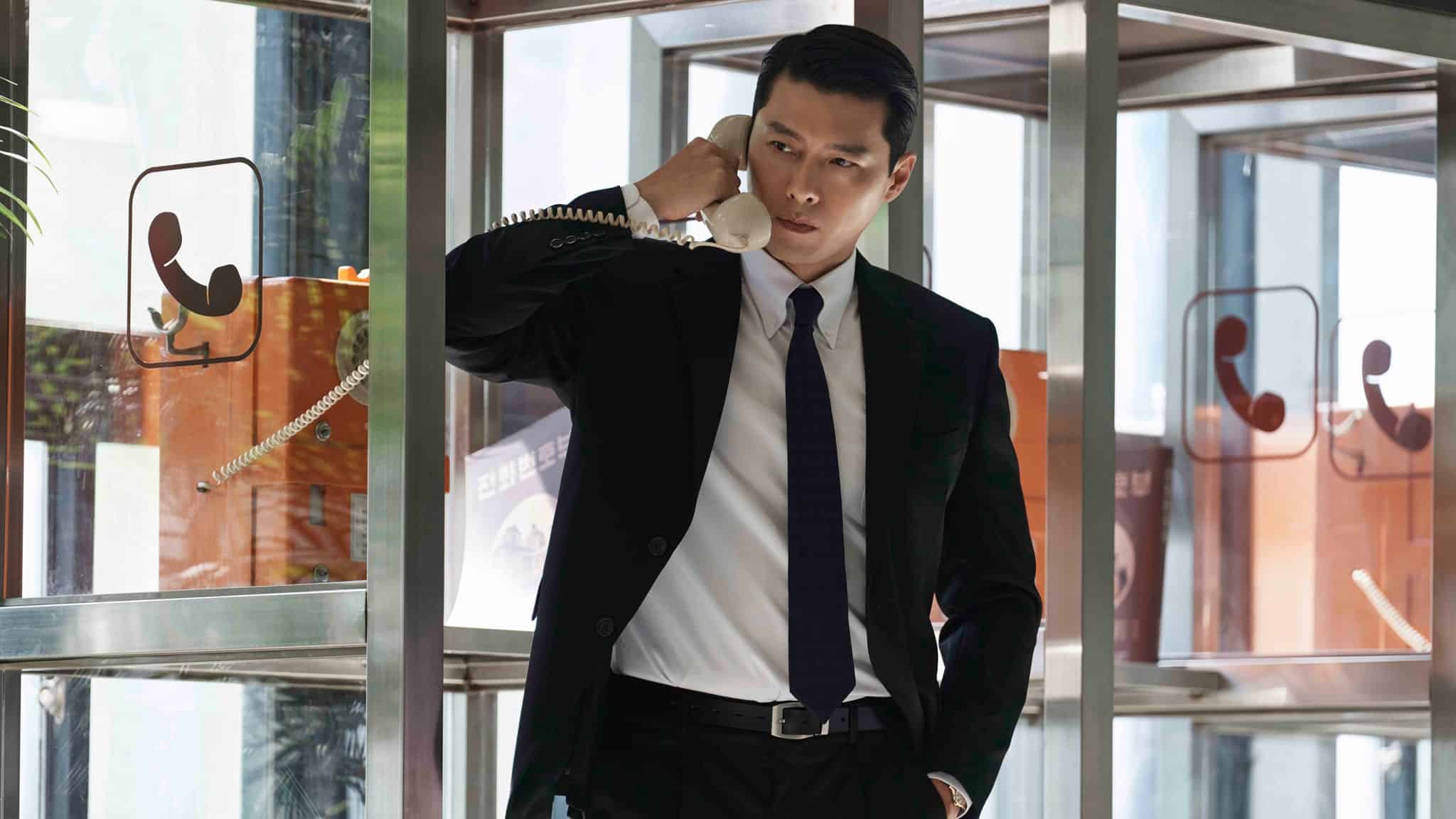 A man in a black suit and tie stands in a glass phone booth, holding a white corded phone to his ear with a serious expression. Orange payphones are visible in the background.