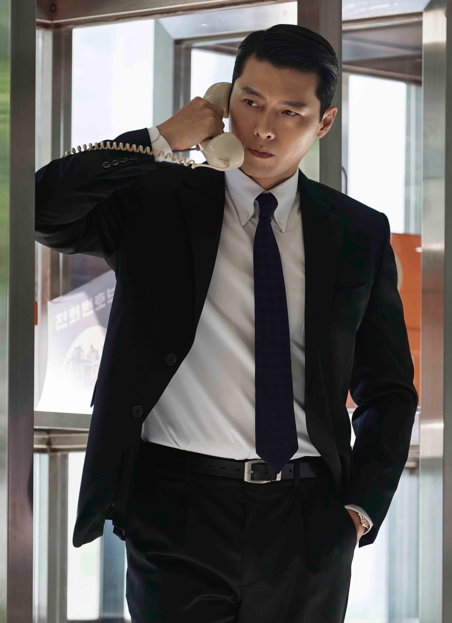 A man in a black suit and tie stands in a glass phone booth, holding a white corded phone to his ear with a serious expression. Orange payphones are visible in the background.