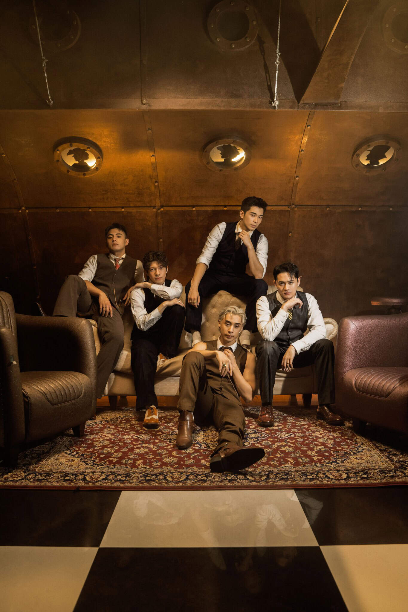 Five men in formal attire pose confidently on and around a vintage sofa in a stylish, dimly lit room with a patterned rug, metallic wall, and black-and-white checkered floor.