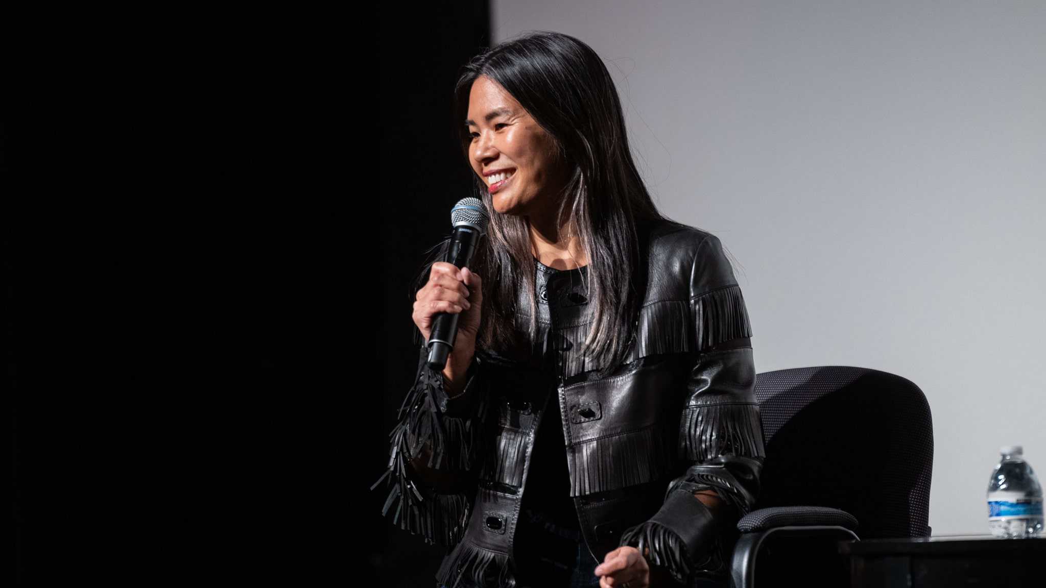 A woman with long dark hair, wearing a black leather jacket with fringe, smiles while speaking into a microphone on stage. She is seated near a table with a bottle of water.