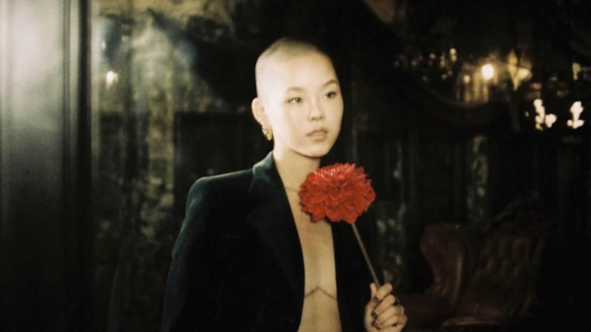 A person with a shaved head, reminiscent of XG’s Cocona, wears a dark velvet blazer and gold earrings while holding a large red flower in a dimly lit, elegant room with vintage decor.