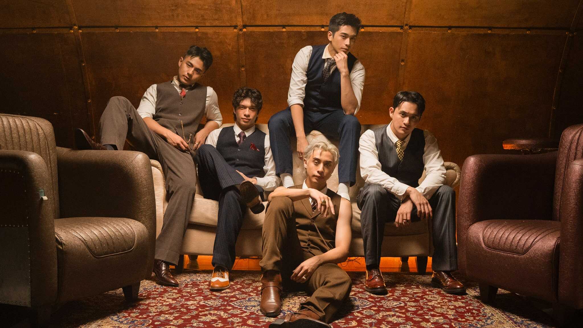 Five young men in vintage suits sit and pose confidently on and around a sofa in a stylish, warm-toned room with leather chairs and a patterned rug, giving a retro, sophisticated vibe.