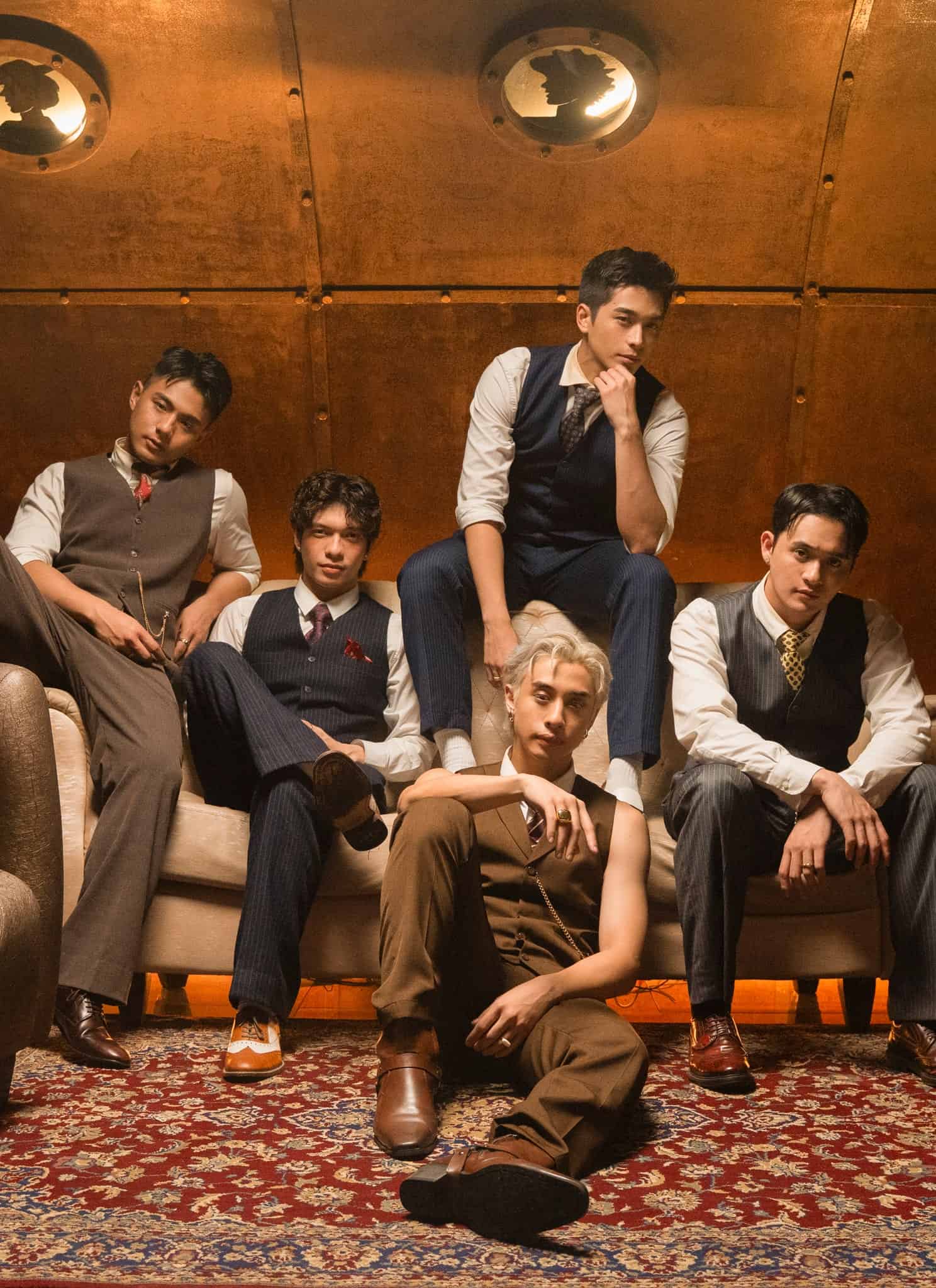 Five young men in vintage suits sit and pose confidently on and around a sofa in a stylish, warm-toned room with leather chairs and a patterned rug, giving a retro, sophisticated vibe.