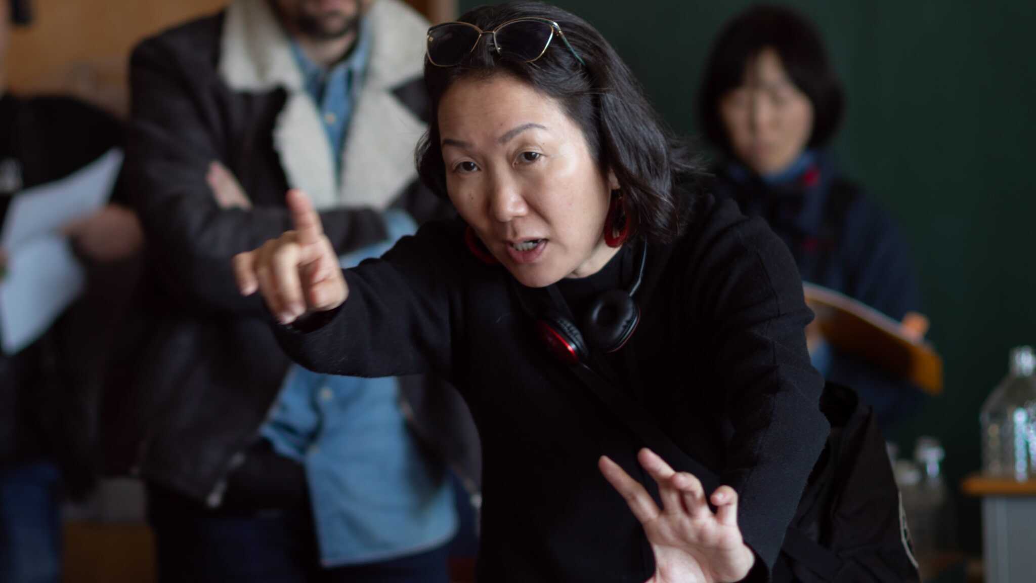 A woman with headphones around her neck gestures with one hand pointing forward and the other held out, speaking intently. Several people are blurred in the background.