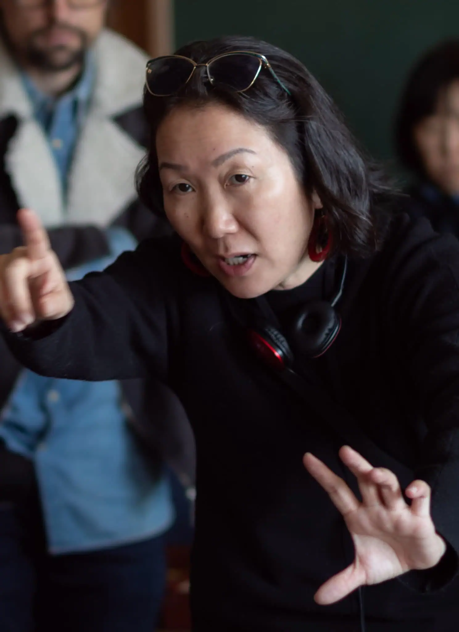 A woman with headphones around her neck gestures with one hand pointing forward and the other held out, speaking intently. Several people are blurred in the background.