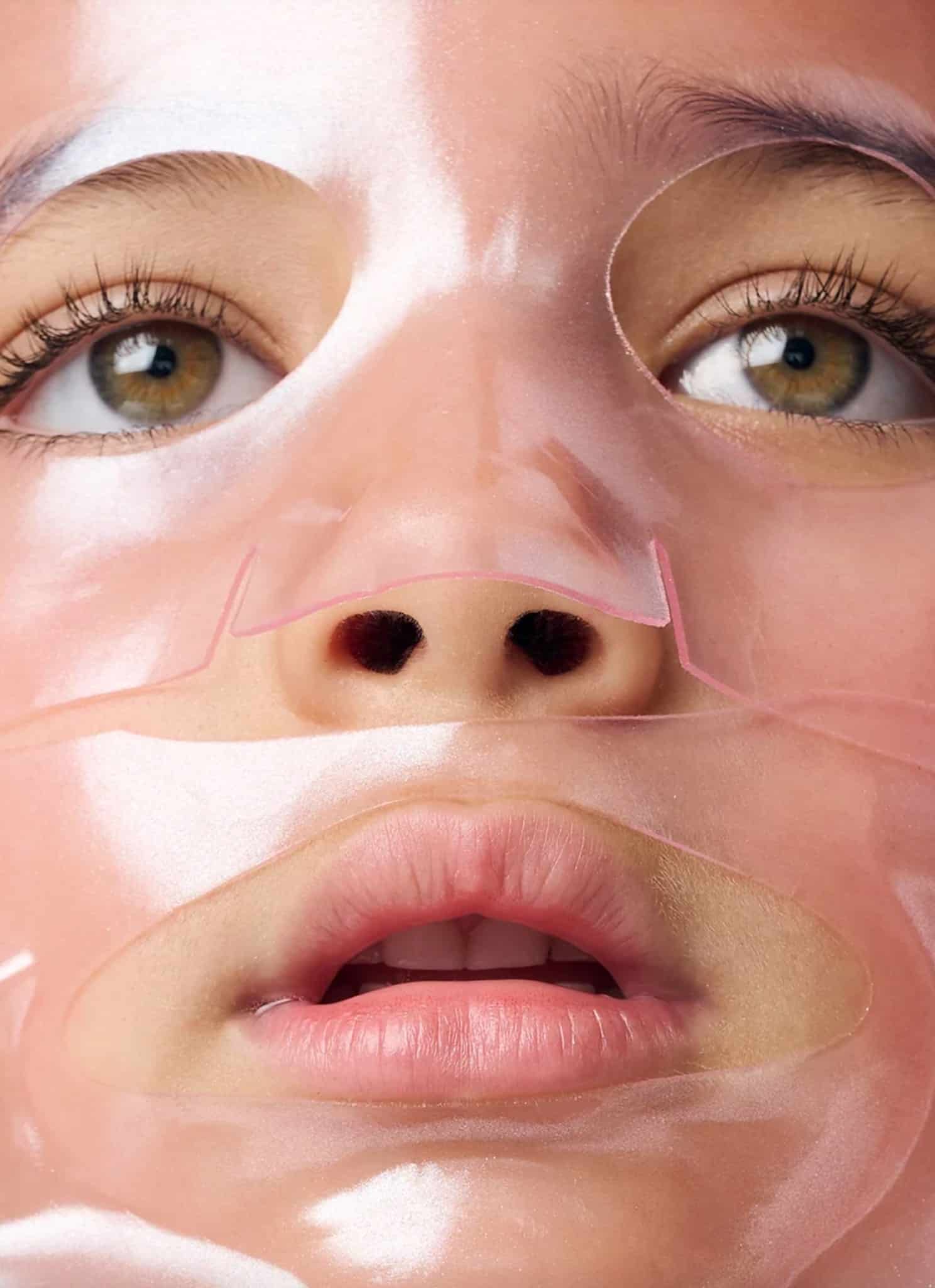 Close-up of a person peeling off a translucent, glossy facial mask, revealing their face underneath. The mask covers most of their face except for the eyes, which are open and looking slightly upward.