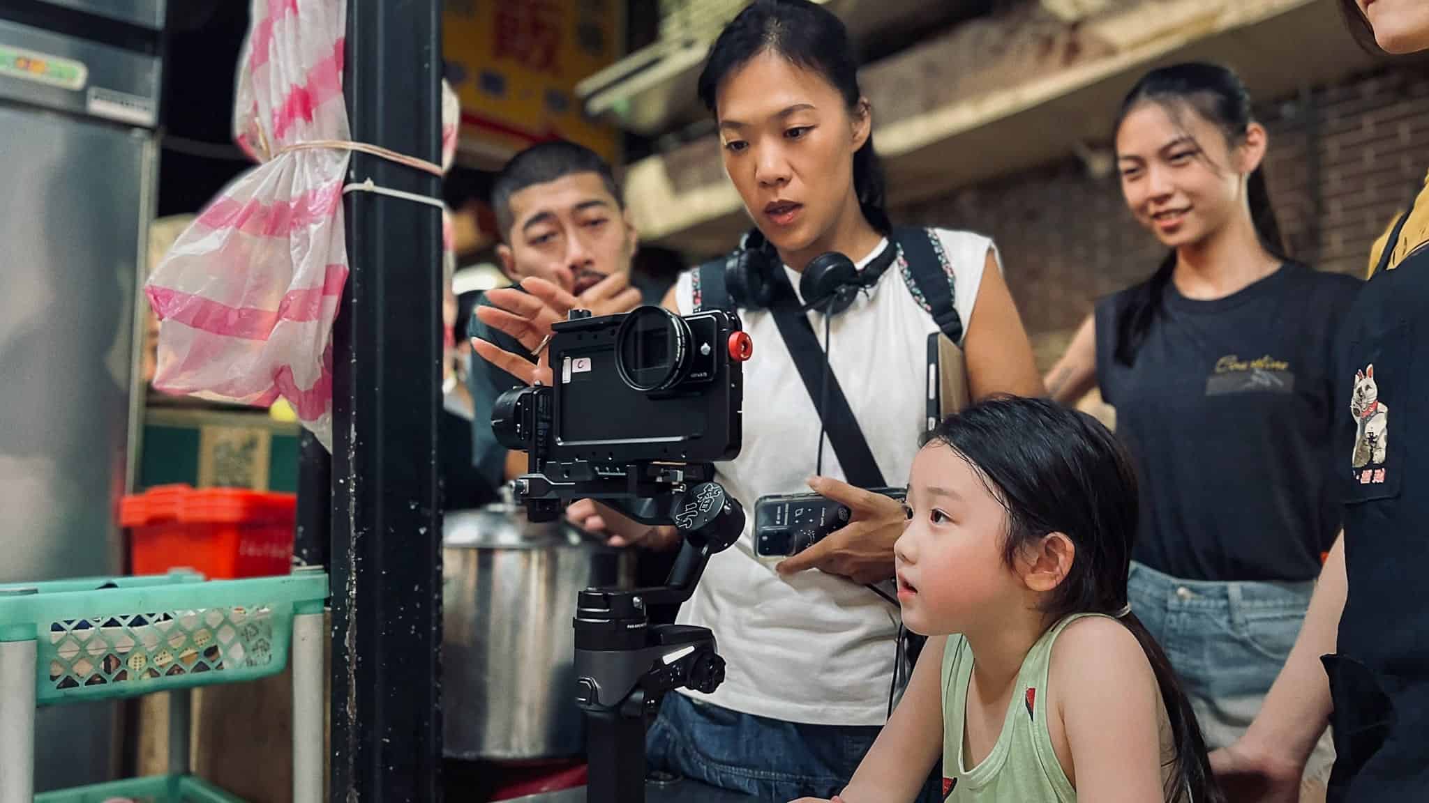 A woman shows a young girl how to use a camera on a tripod, while three other adults look on. They appear to be in an indoor market or food stall setting.