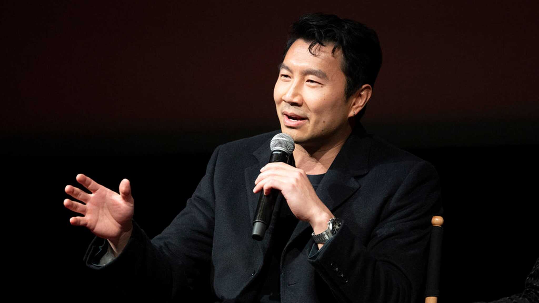 A person sits on a chair, holding a microphone and speaking. They are wearing a dark jacket and gesturing with one hand. The background is dark.