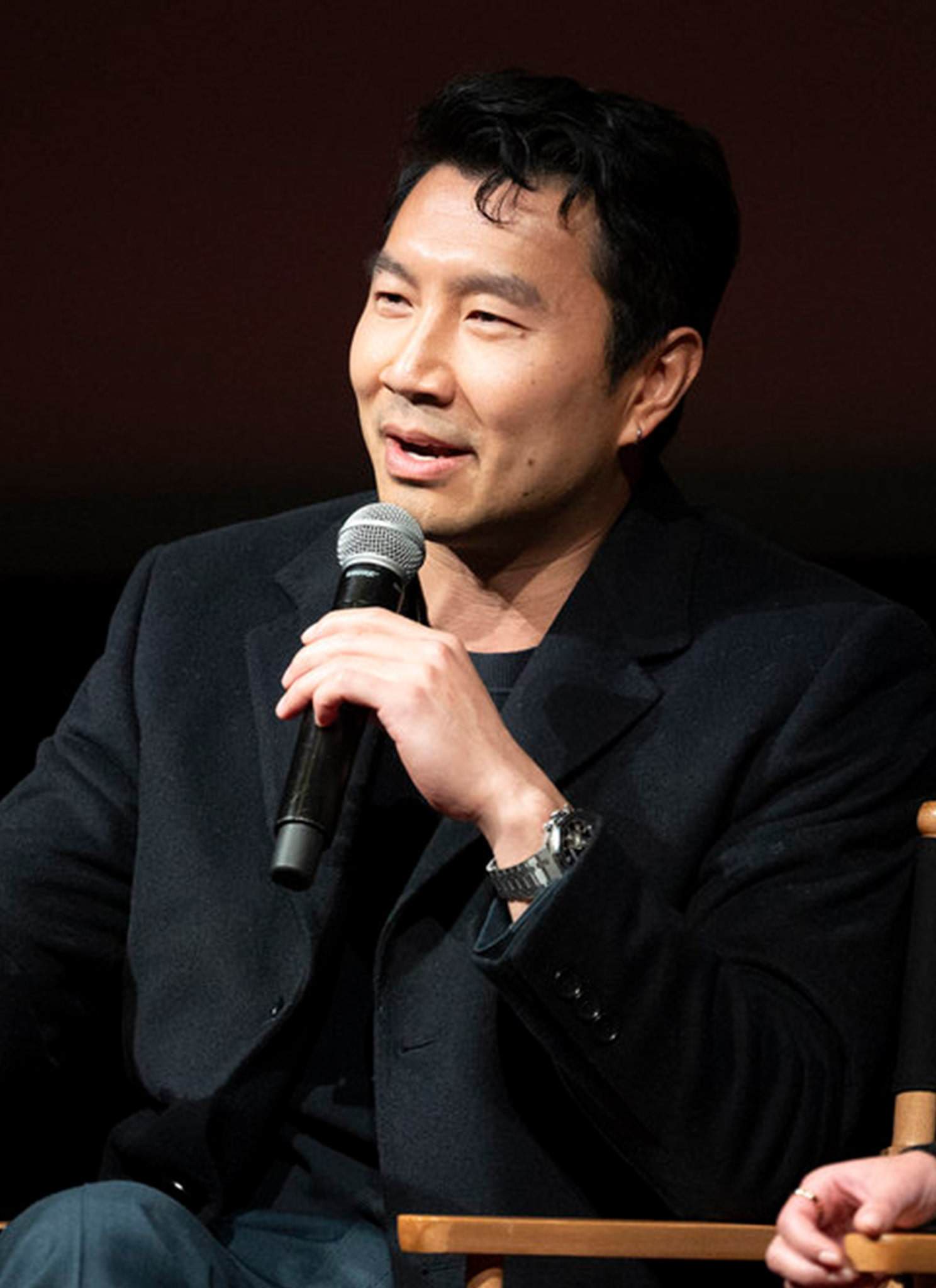 A person sits on a chair, holding a microphone and speaking. They are wearing a dark jacket and gesturing with one hand. The background is dark.