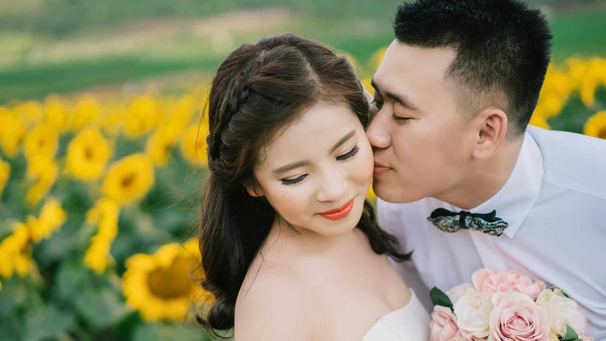 A groom in a white shirt kisses a brides cheek as she smiles, holding a pink bouquet. They stand close together in a vibrant sunflower field.