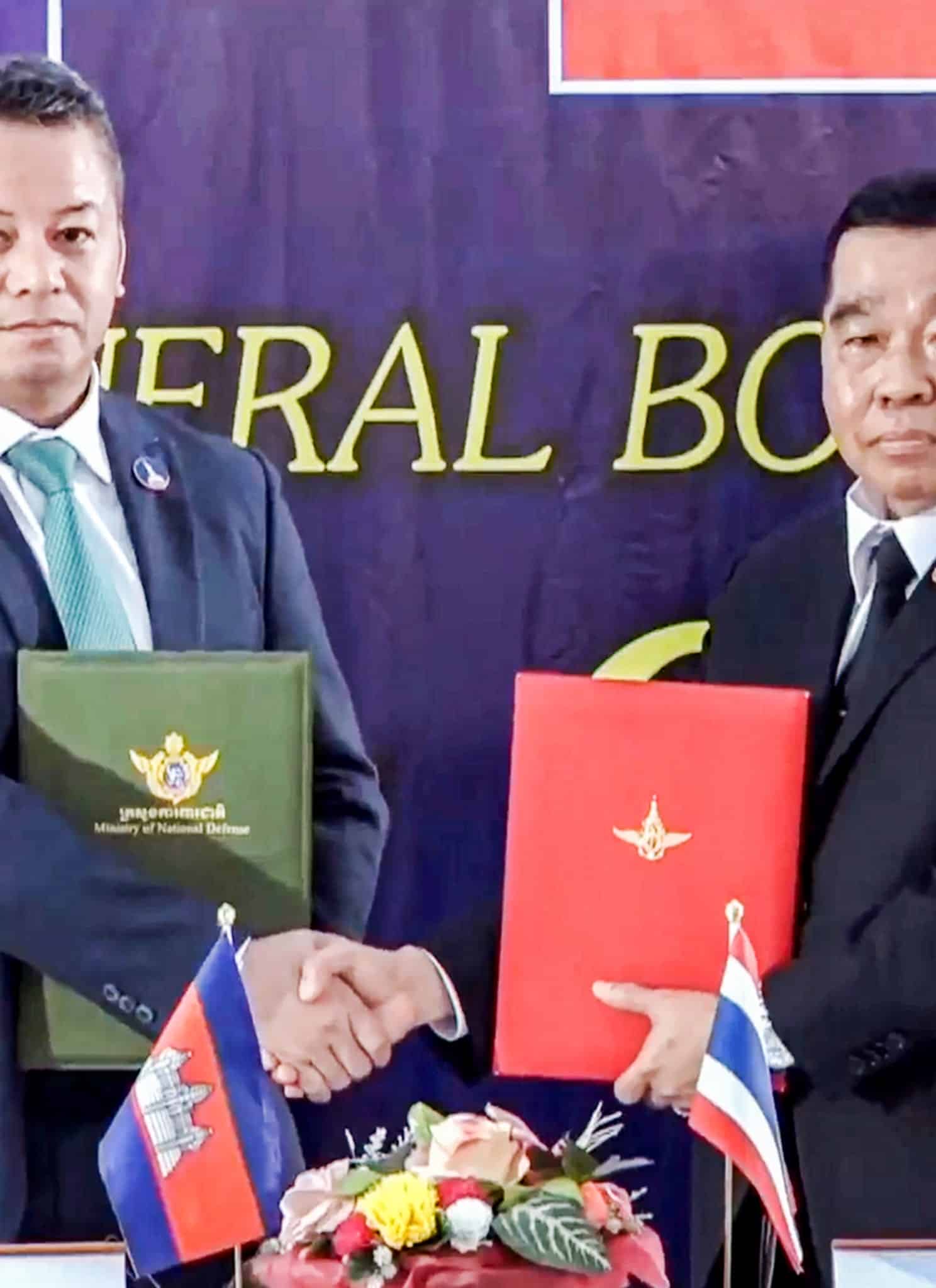 Two men in suits shake hands while holding official folders, one green and one red, in front of a backdrop with English text—an apparent step toward resolving the Thailand Cambodia conflict. Both sit at a table with microphones, water glasses, and a Cambodian flag visible.