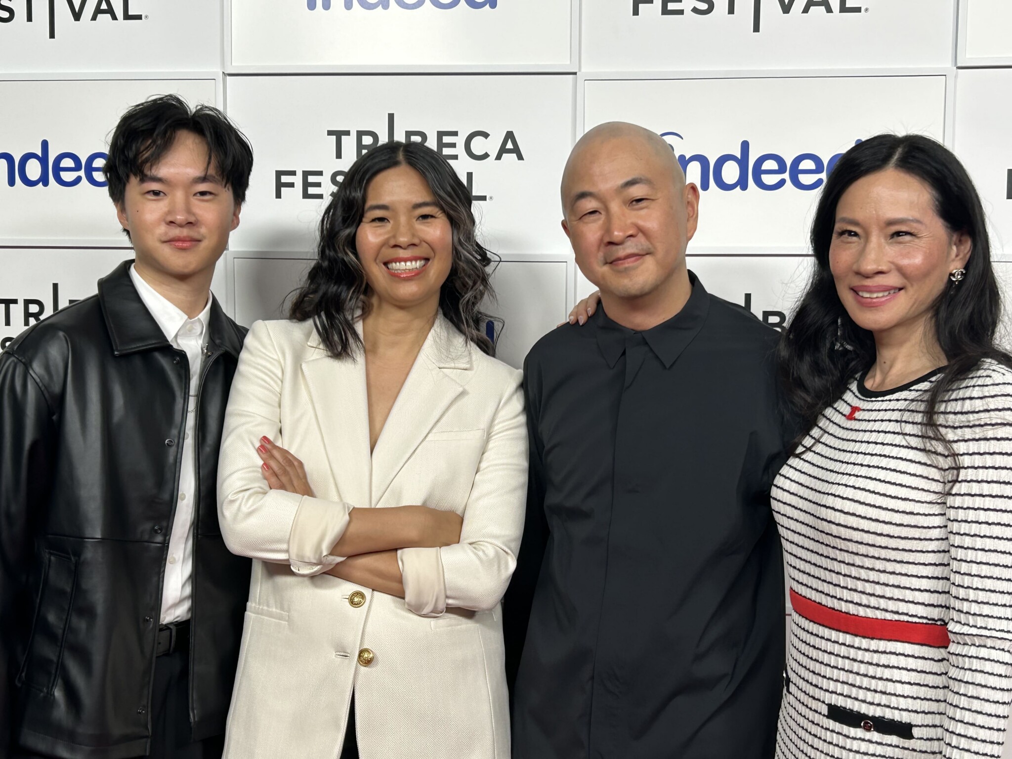 Four people stand smiling together in front of a photo backdrop with “Tribeca Festival” and sponsor logos. They are dressed in semi-formal to stylish attire and are posing for the camera.