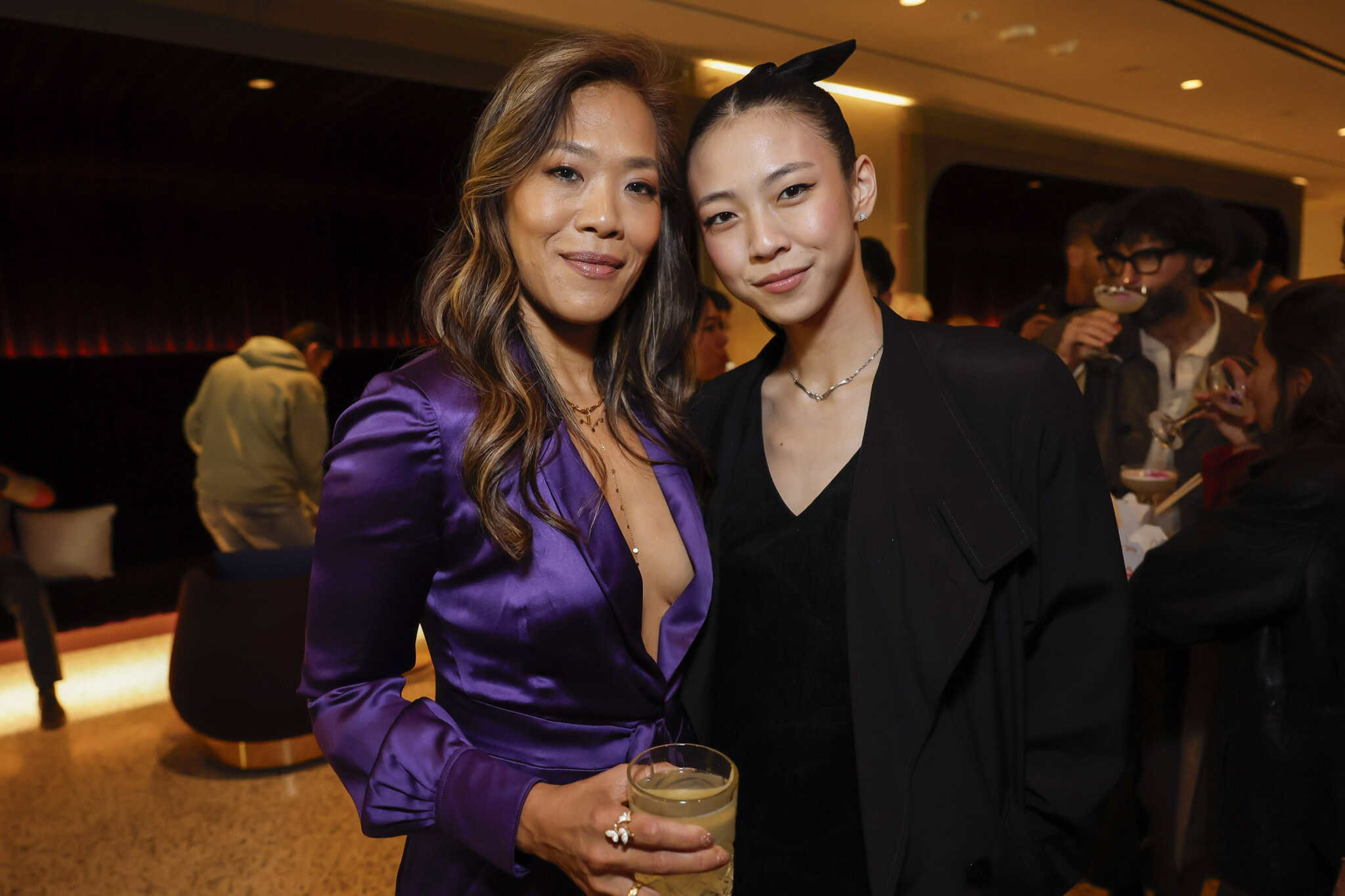 Two women pose together at an indoor social event; one wears a shiny purple dress and holds a drink, while the other is dressed in black. People and soft lighting are visible in the background.
