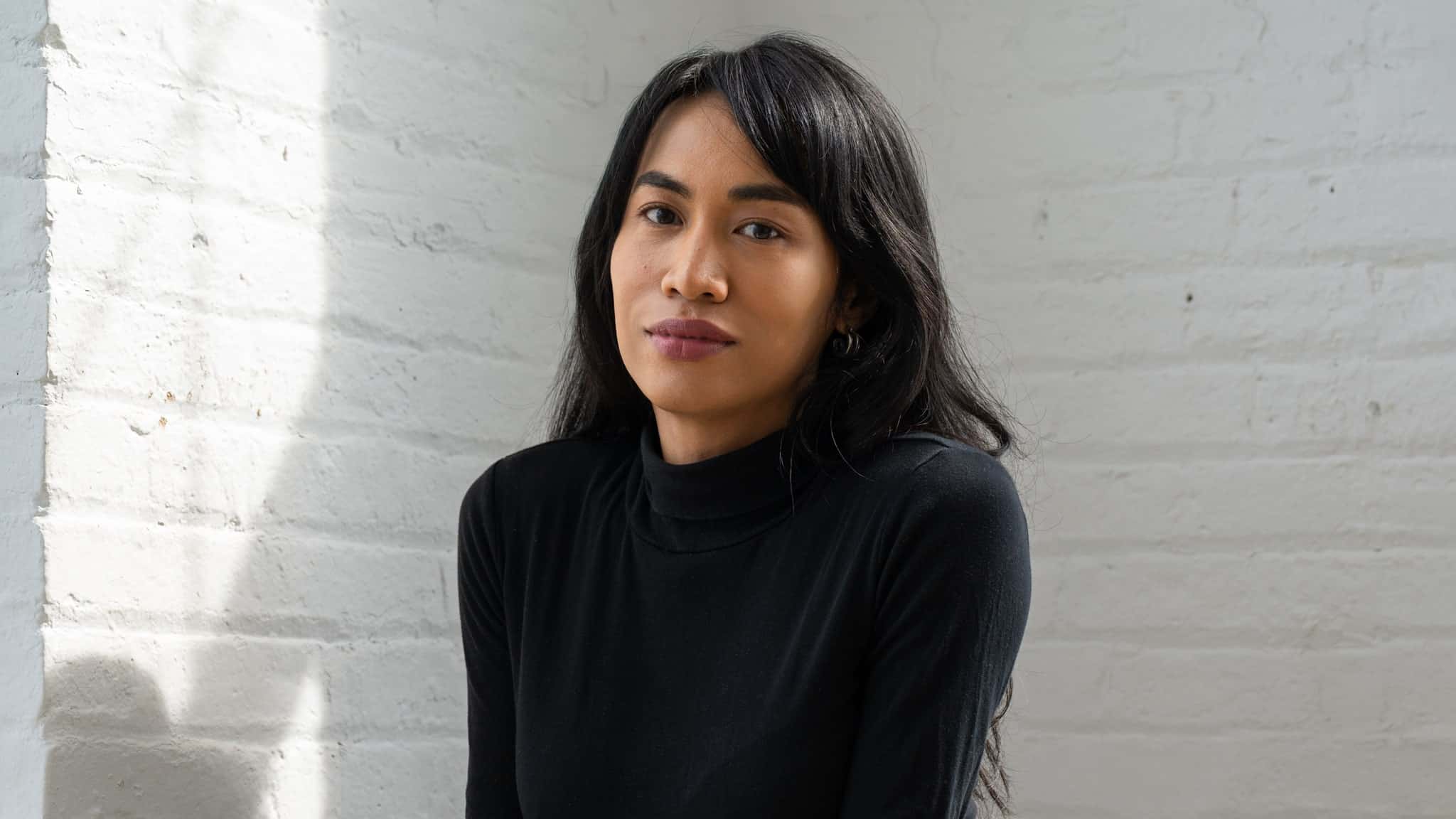 A woman with long dark hair wearing a black turtleneck sits against a white brick wall, softly lit by sunlight coming from the left. She looks calmly at the camera with a neutral expression.