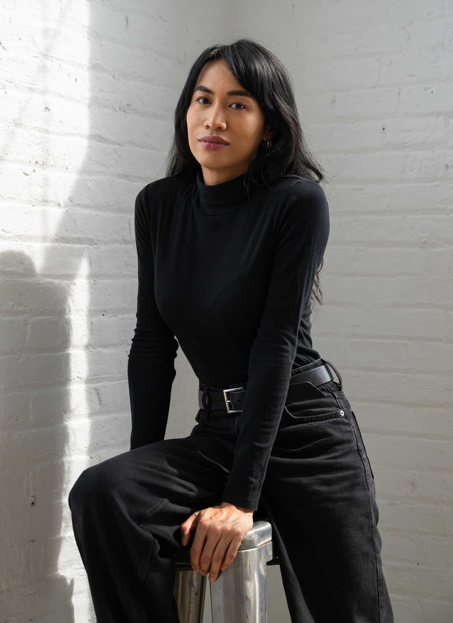 A woman with long dark hair wearing a black turtleneck sits against a white brick wall, softly lit by sunlight coming from the left. She looks calmly at the camera with a neutral expression.