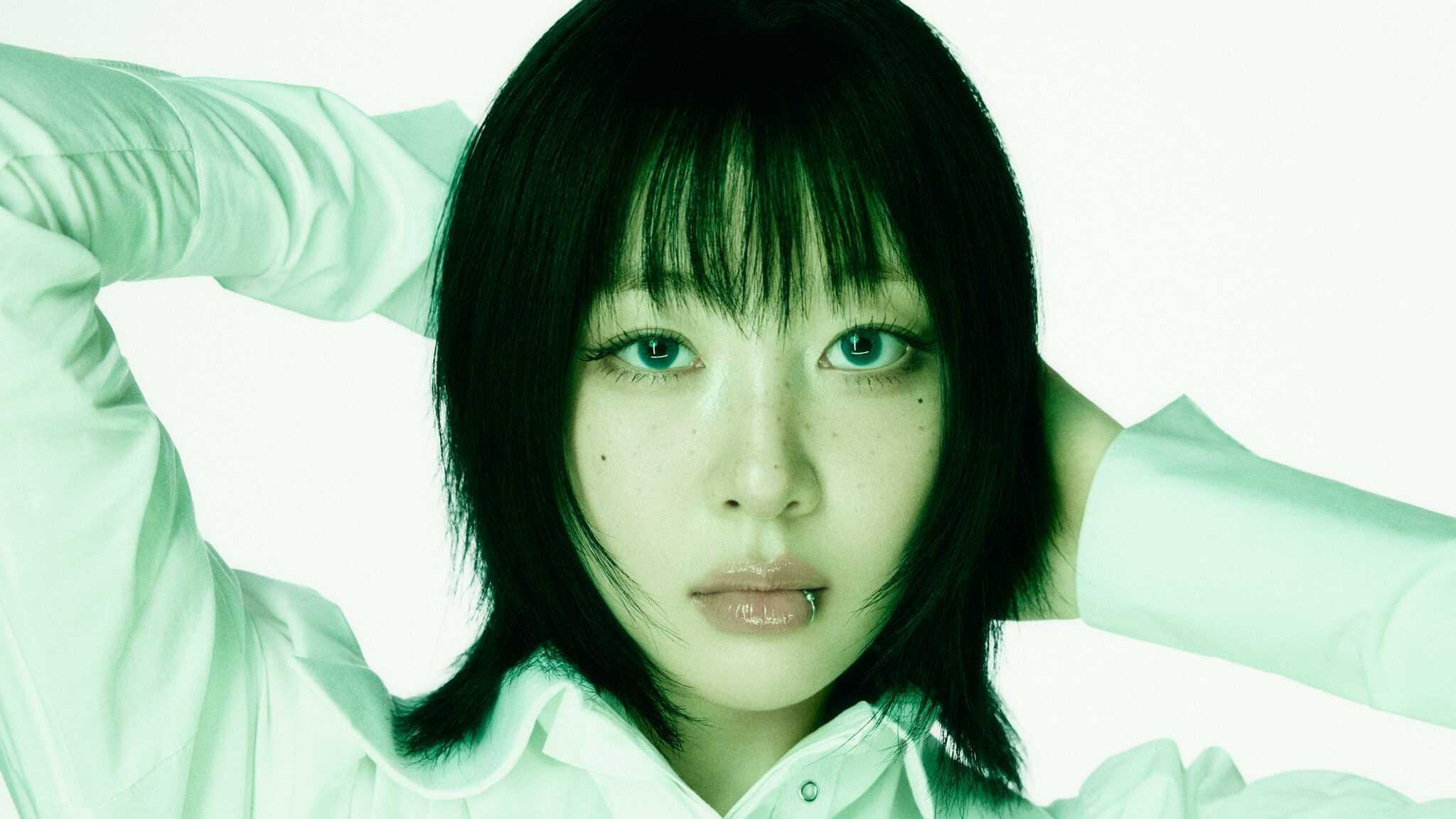 A person with straight, shoulder-length black hair and bangs poses with hands behind their head. They have light makeup, glossy lips, a piercing on the lower lip, and wear a white collared shirt against a light green background.