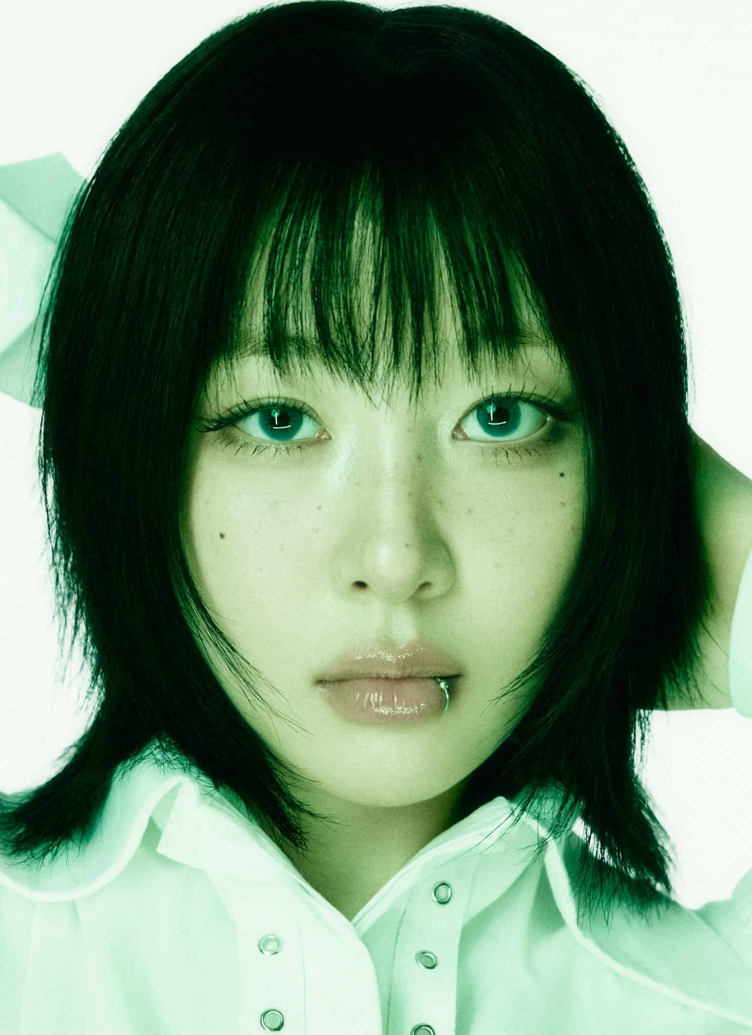 A person with straight, shoulder-length black hair and bangs poses with hands behind their head. They have light makeup, glossy lips, a piercing on the lower lip, and wear a white collared shirt against a light green background.