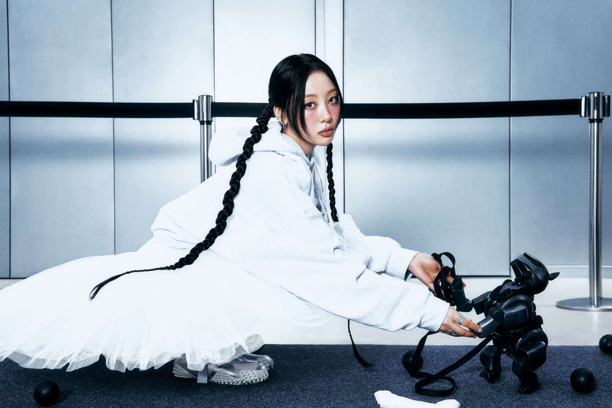 A woman in a white hoodie and tutu crouches on the floor, holding the paw of a black robotic dog. She has long braids and is surrounded by scattered black balls, with a gray wall and stanchions in the background.