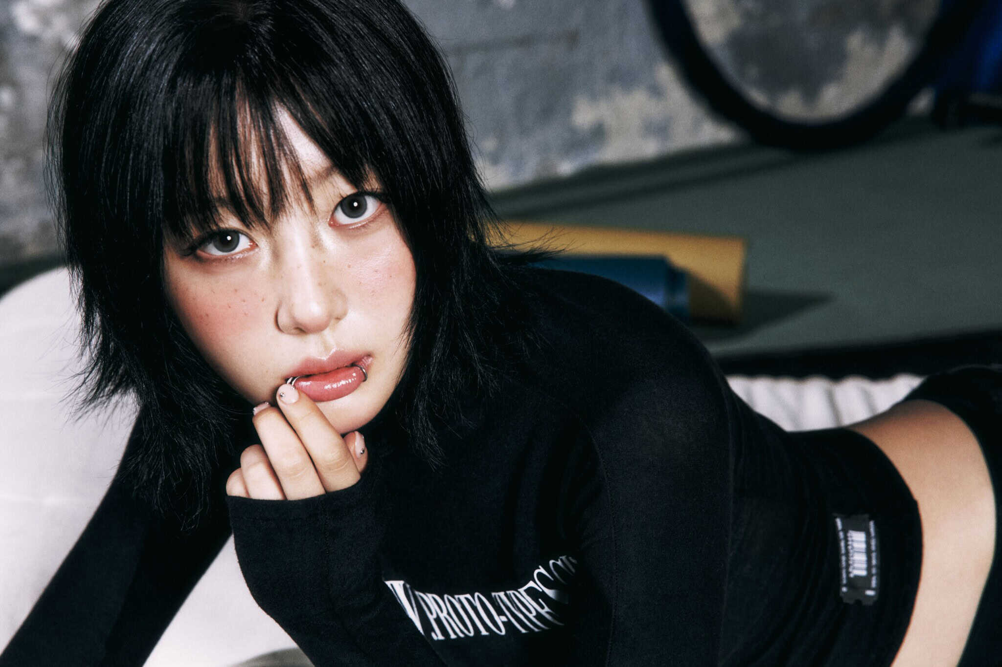 A person with short, dark hair and bangs wears a black long-sleeve top, looking directly at the camera with a thoughtful expression and a finger near their lips. The background is blurred and industrial.